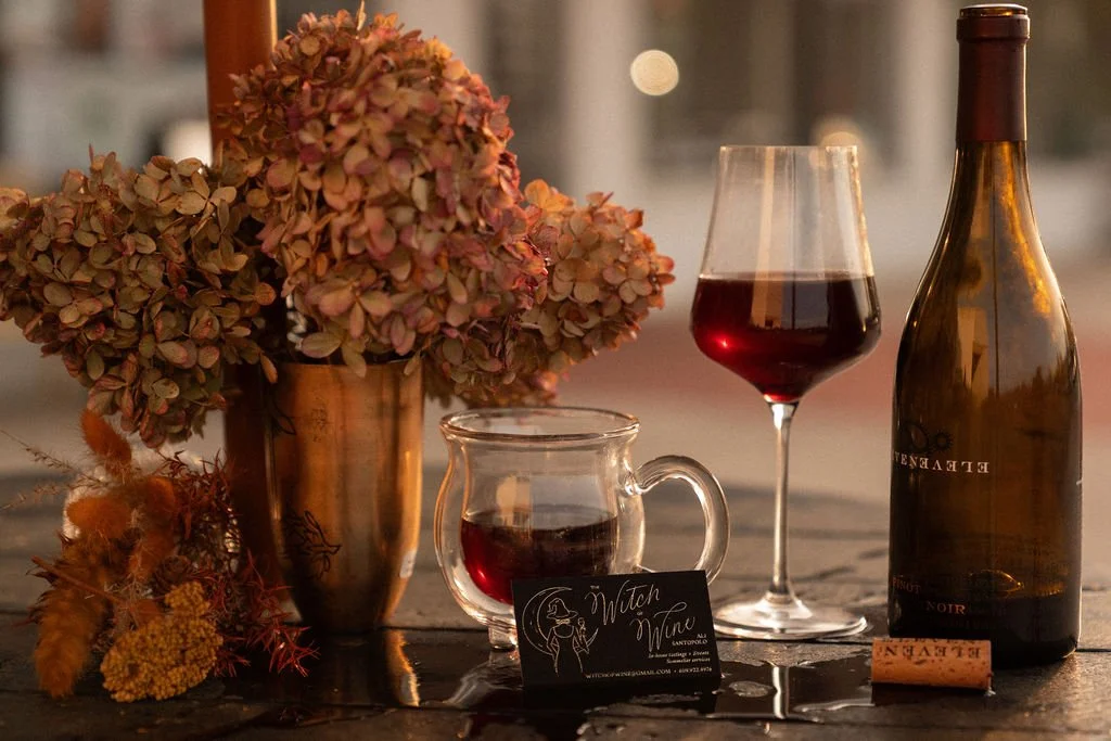 A table with a vase of dried pink hydrangeas, a glass of red wine, a small pitcher of red liquid, a bottle of red wine, and a wine cork, with a small black business card for 'Witch of Wine' placed in front of the pitcher.