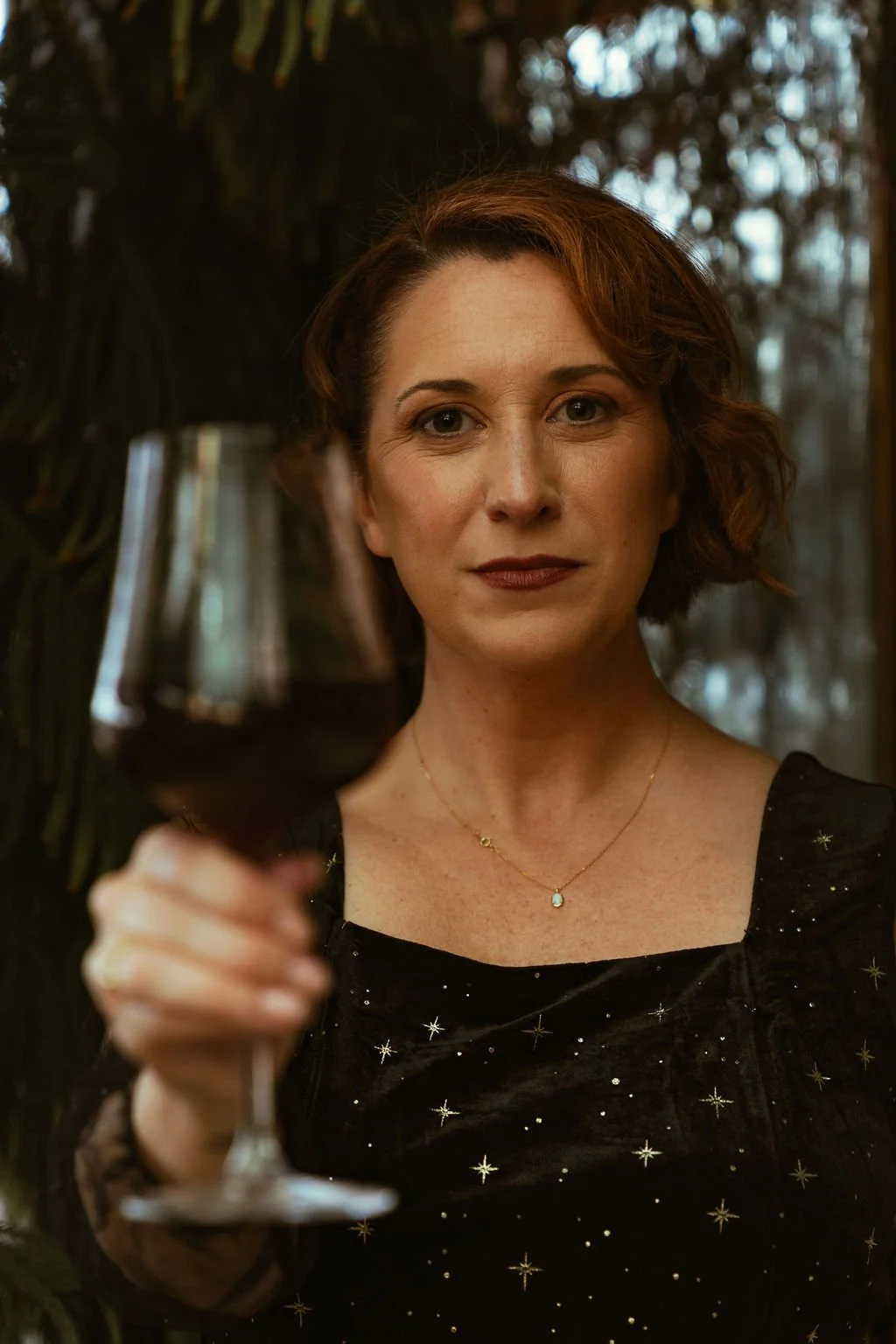 A woman with short, wavy, reddish-brown hair looks directly at the camera while holding a glass of red wine. She is wearing a black dress with star patterns and a delicate gold necklace, standing in a cozy, dimly-lit environment with greenery in the background.