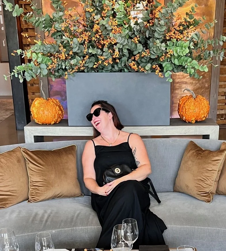 Woman in black dress and sunglasses sitting on a sofa, laughing, with decorative orange pumpkins and large leafy green and orange floral arrangement in the background.