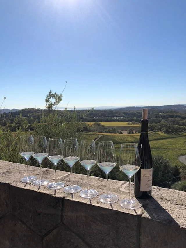 Seven wine glasses and a bottle of wine on a stone wall overlooking a scenic countryside landscape with trees and fields under a clear blue sky.