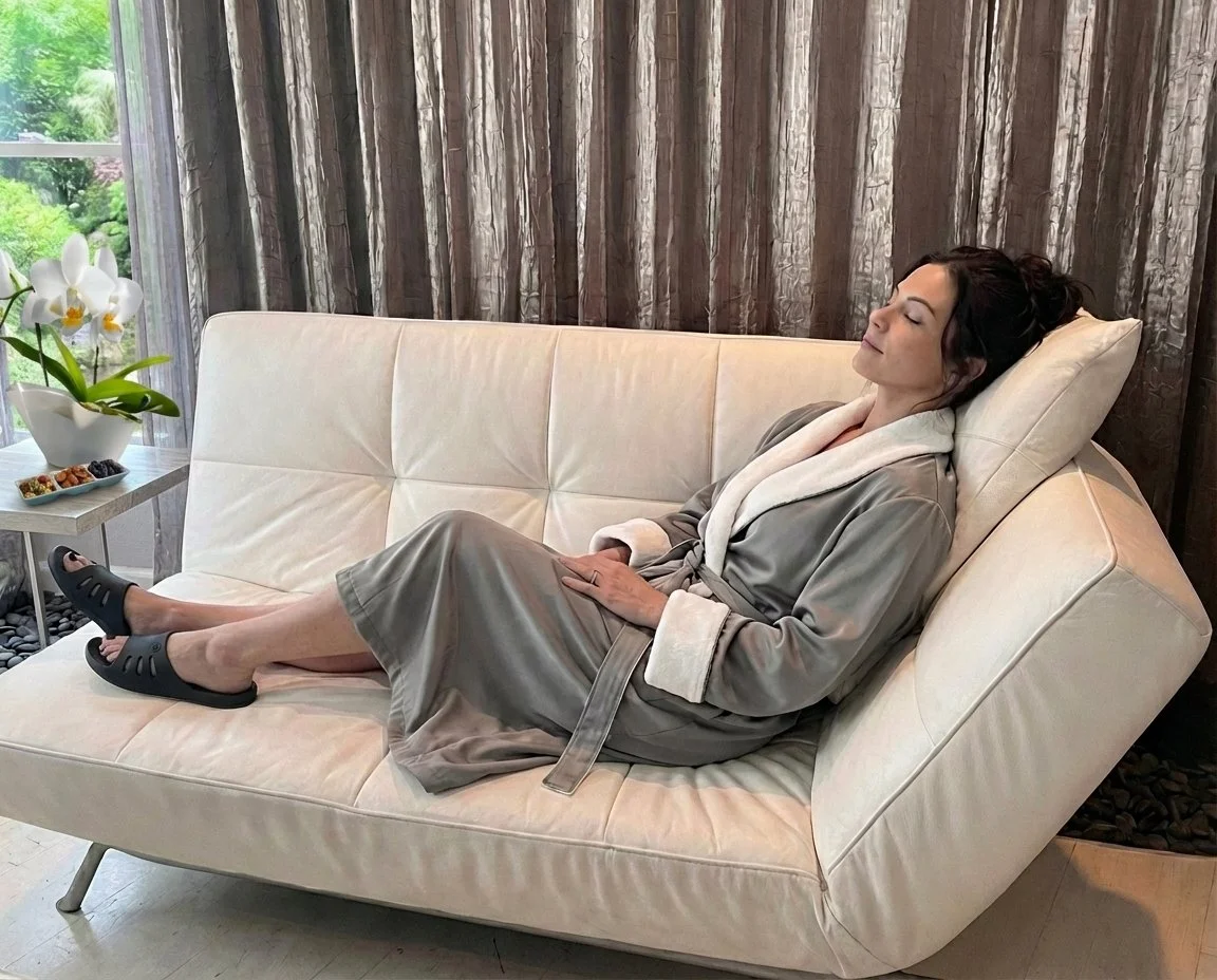 A woman relaxing on a cream-colored sofa with her eyes closed, wearing a gray satin robe, black slippers, and resting her head on a beige pillow against a dark curtain backdrop, with a window and a side table with a white orchid and snacks nearby.