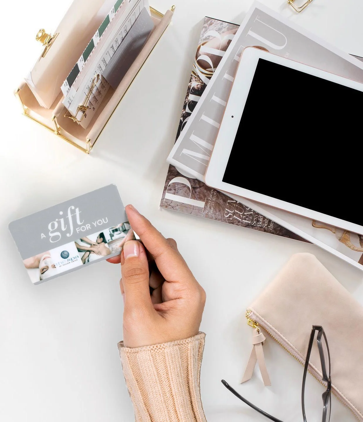 A person's hand holding a gift card in front of a white desk. On the desk are magazines, an iPad, a beige zippered pouch, and a pair of eyeglasses. There is a gold wire basket with files and papers in the background.