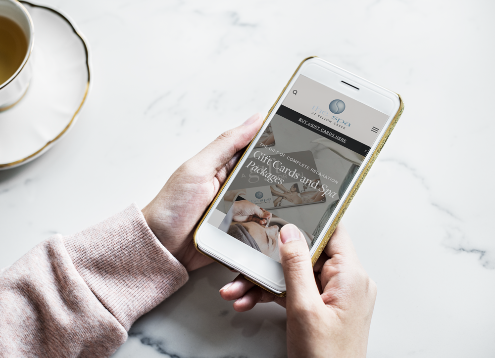 Person holding a smartphone displaying a website for gift cards and spa packages, with a tea cup on a saucer nearby on a white marble surface.
