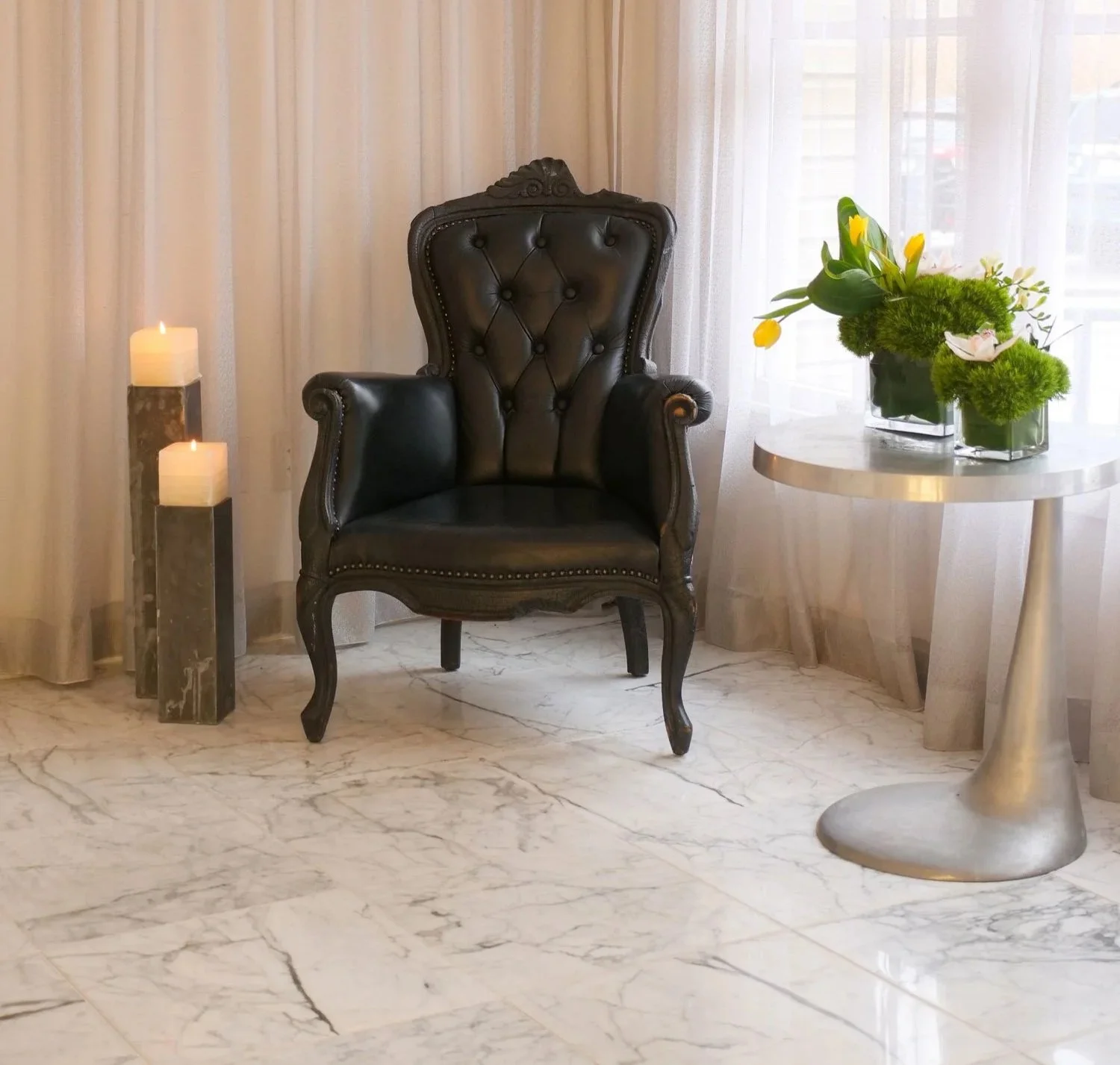 A black leather armchair with a tall, tufted backrest and carved wooden frame, positioned in a cozy interior space. To the left of the chair are two lit candles on a marble stand, and to the right, a round table with a glass vase containing yellow tu