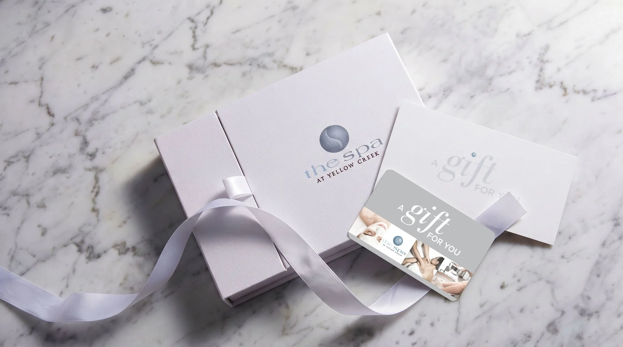 A white gift box with a satin ribbon, placed on a marble surface. The box has the logo and name 'the spa at Yellow Creek' on it. Next to the box is a gift card with similar branding, featuring images of a woman receiving a spa treatment.