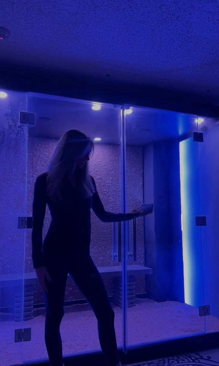 A woman standing inside a room with blue ambient lighting, near glass walls and a metal structure, with a glowing blue vertical light on the right.