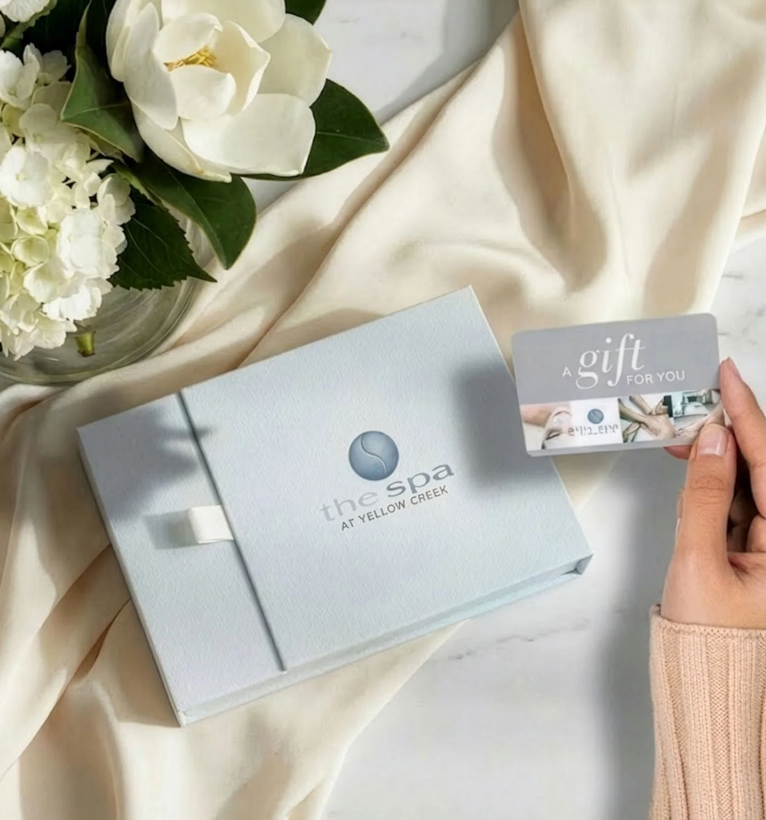 A white box with a logo and text for 'the spa at yellow creek' is on a cream-colored fabric. A hand holds a gift card that says 'a gift for you' while a bouquet of white flowers sits nearby.