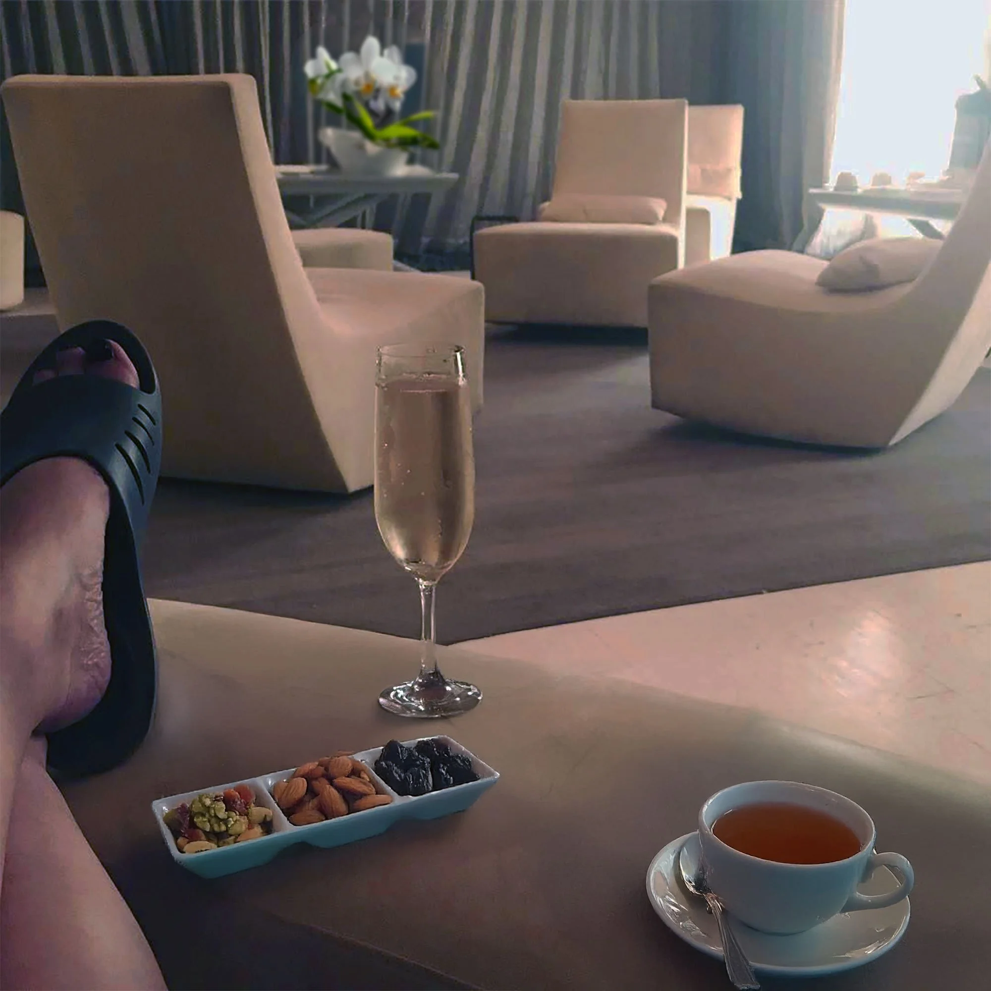 Tranquility Lounge with cream colored chairs, a glass of champagne, a cup of tea, and a plate of snacks on a table, with a person wearing a grey spa sandals relaxing in the foreground.