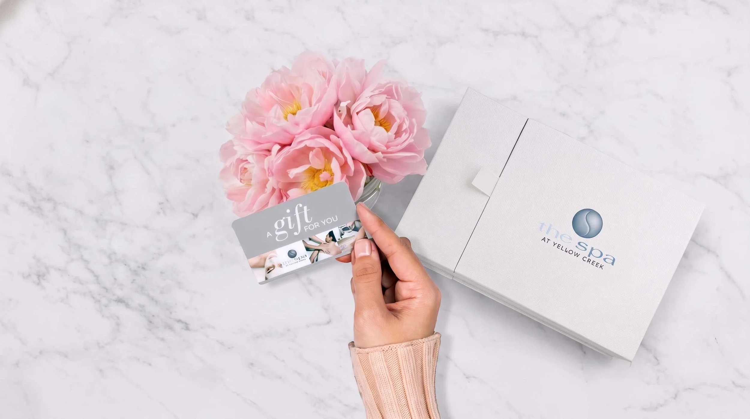 A person holding a gift card with a bouquet of pink flowers nearby, on a marble surface; a white box from The Spa at Yellow Creek is also visible.