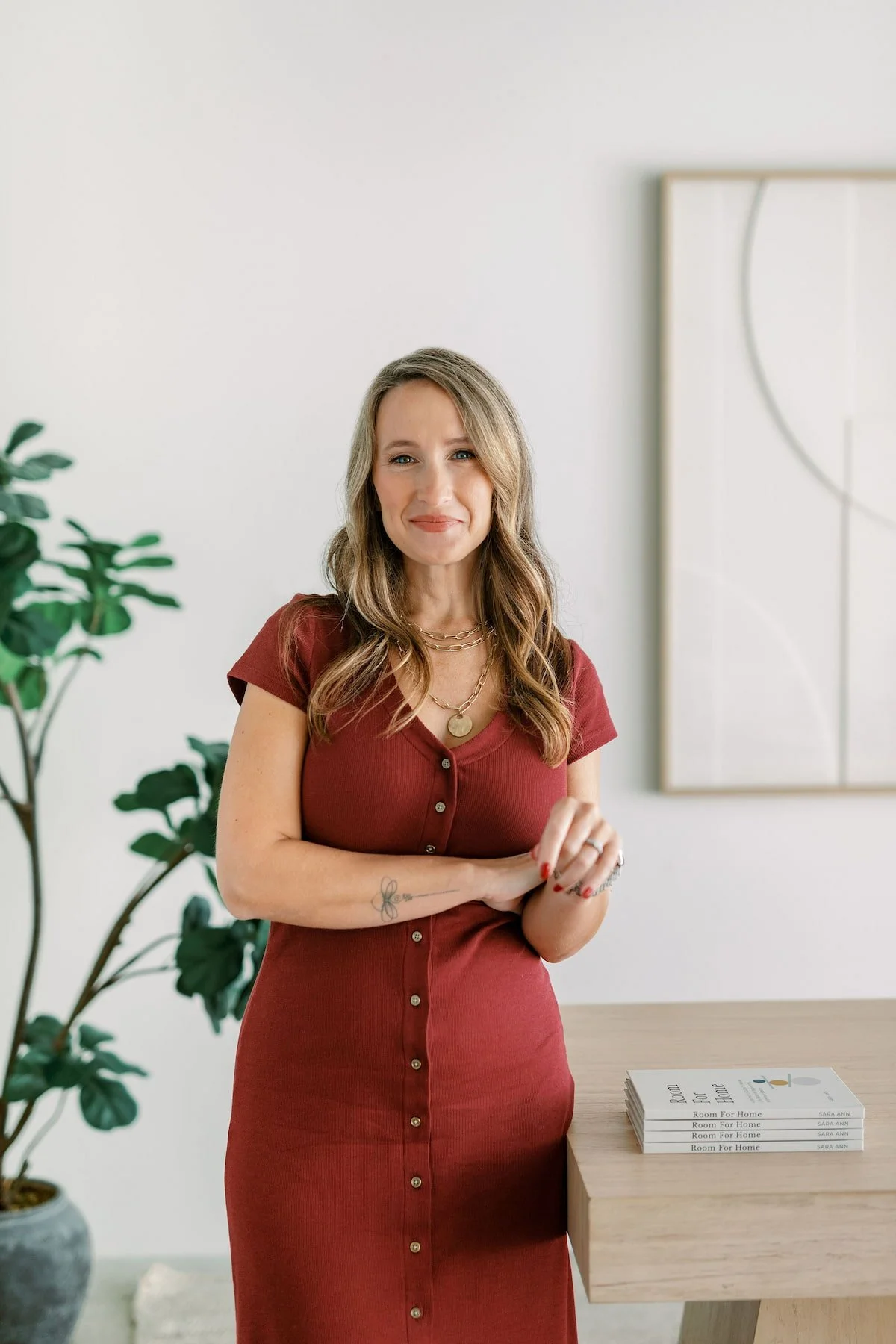 A woman with wavy light brown hair, wearing a red button-up dress, standing in a room with a white wall, a potted plant, and a table with books.