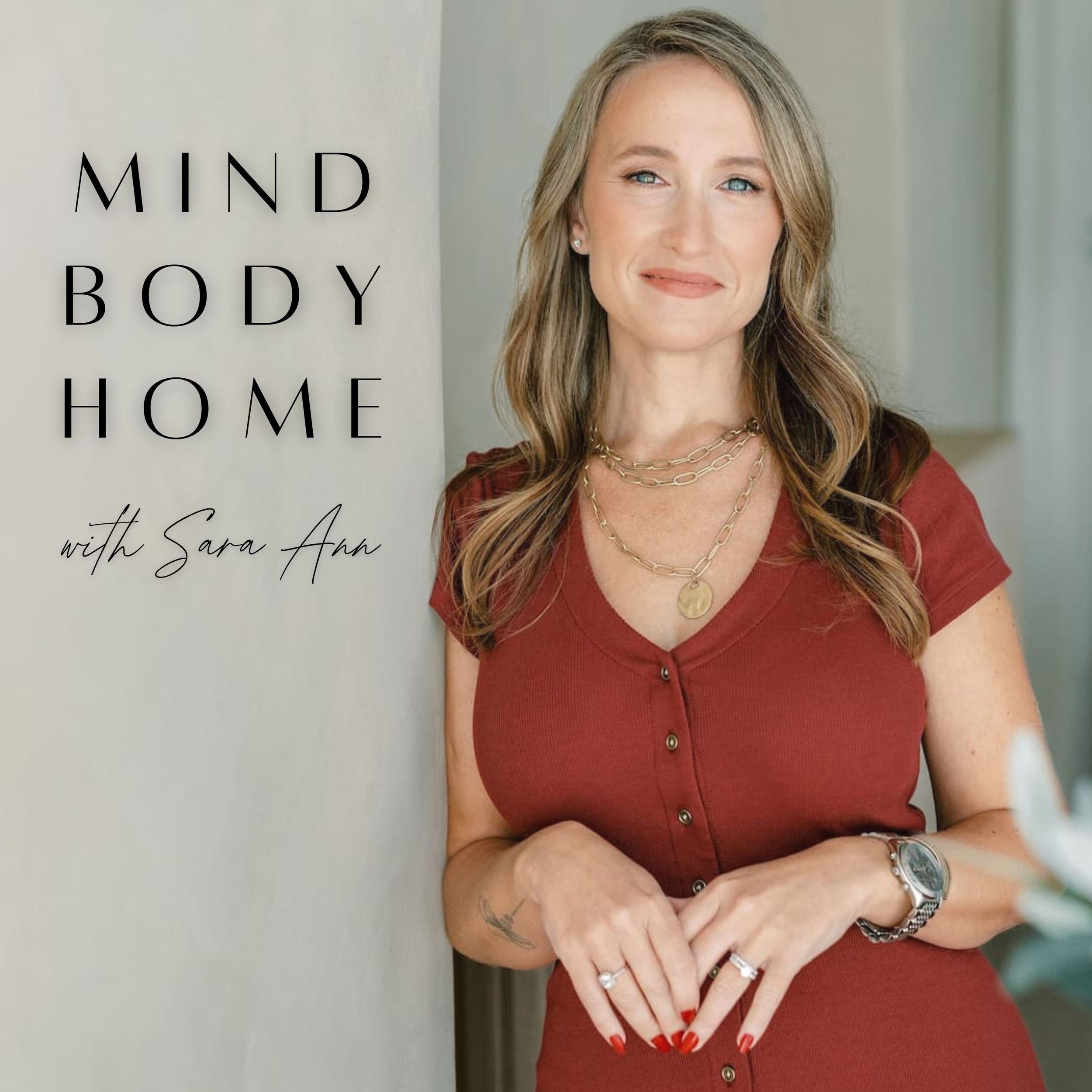 A woman with long, wavy, light brown hair and blue eyes, wearing a red short-sleeved button-up top, gold necklaces, a watch, and rings, standing next to a light-colored wall with a podcast title 'Mind Body Home with Sara Ann' in black and cursive text.