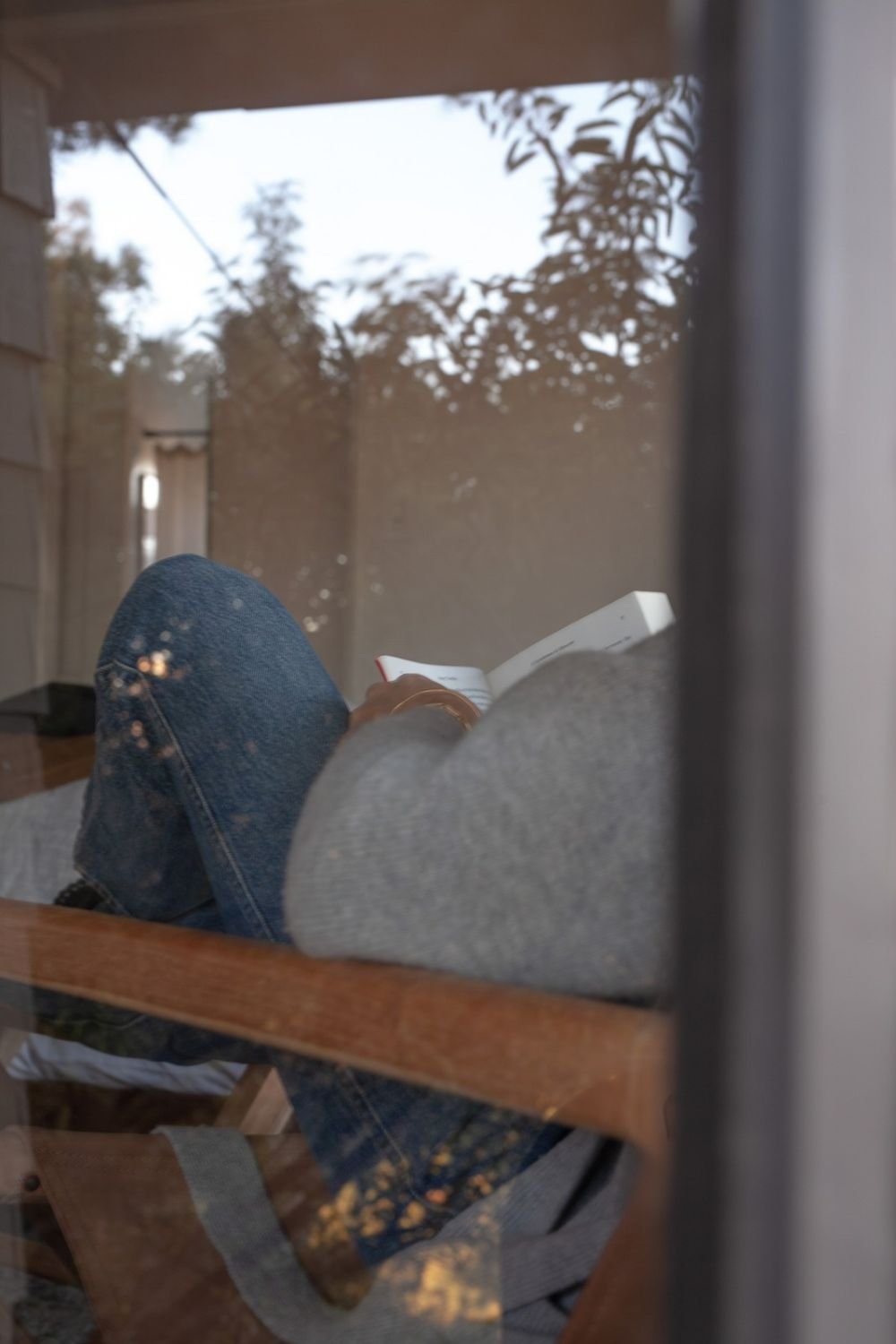 Shot looking into window. Woman in blue jeans and light grey sweater sitting in a chair with wooden arms reading a book.