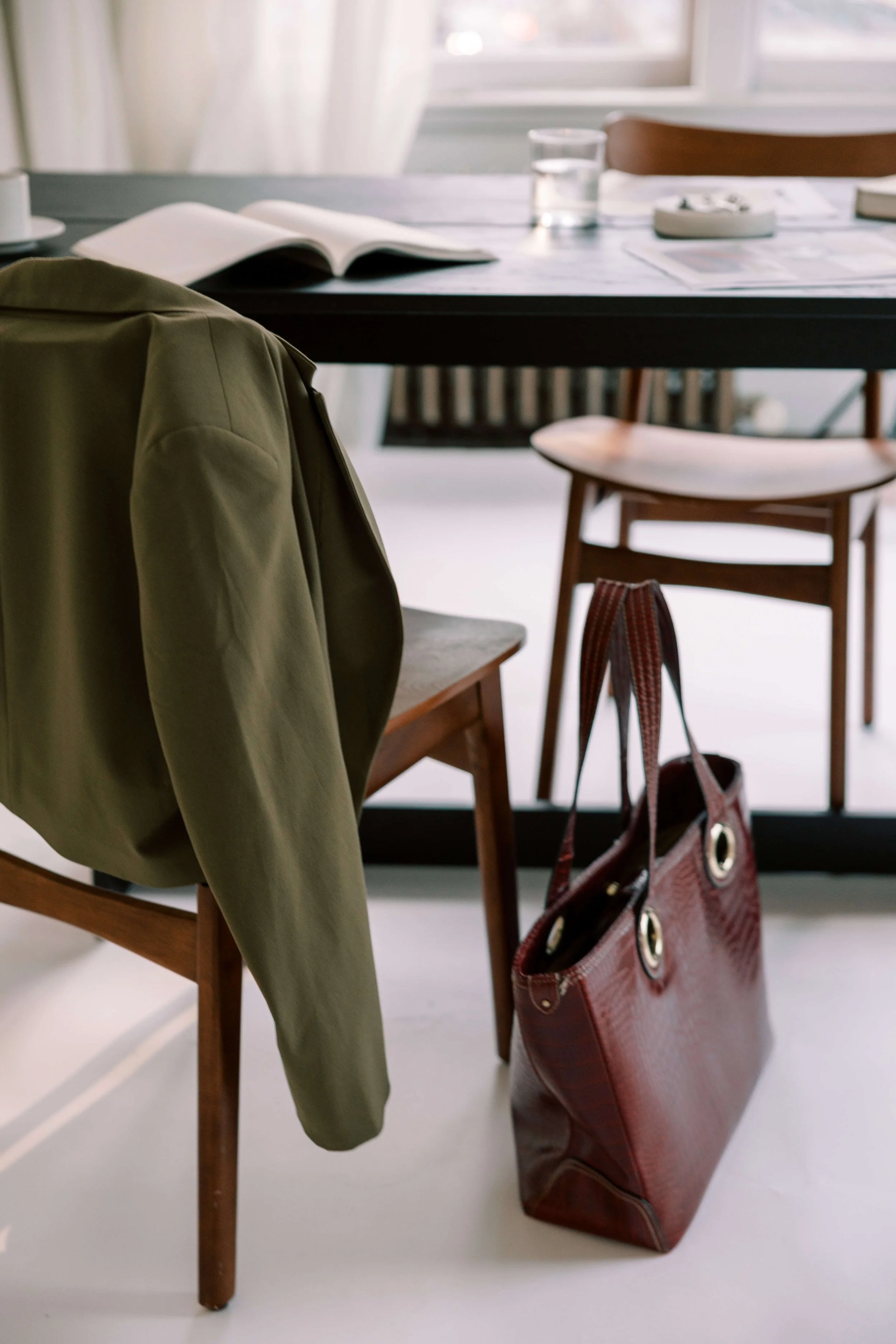 An interior scene featuring a wooden chair with a green jacket hanging on it, a red handbag placed on the floor, and a black table with an open book, a glass of water, and some papers.