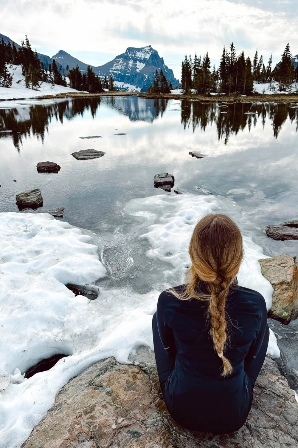 Woman with long blonde braid and blue shirts sits atop of a large rock looking out onto the frozen lake cover withe snowy mountains ahead.