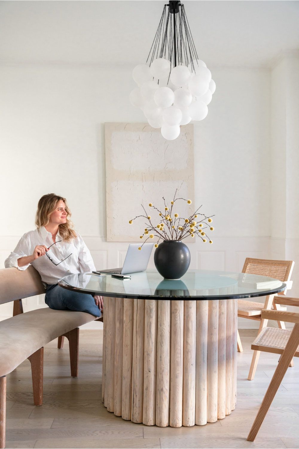 Woman sitting at a modern round dining table with a laptop in a bright, minimalist home office and dining space featuring natural wood textures, neutral tones, and sculptural lighting.
