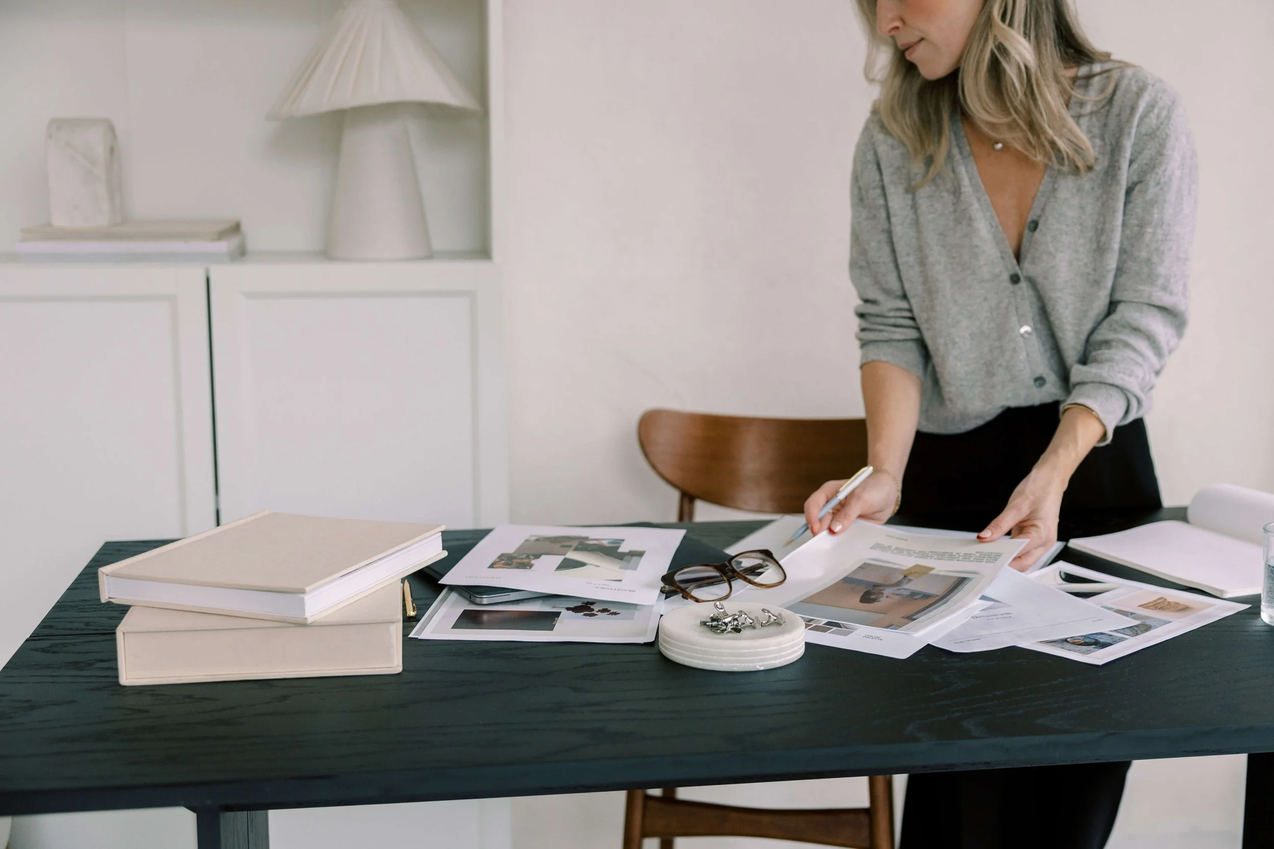 Woman leaning over a table covered with papers, photographs, and a pair of glasses.