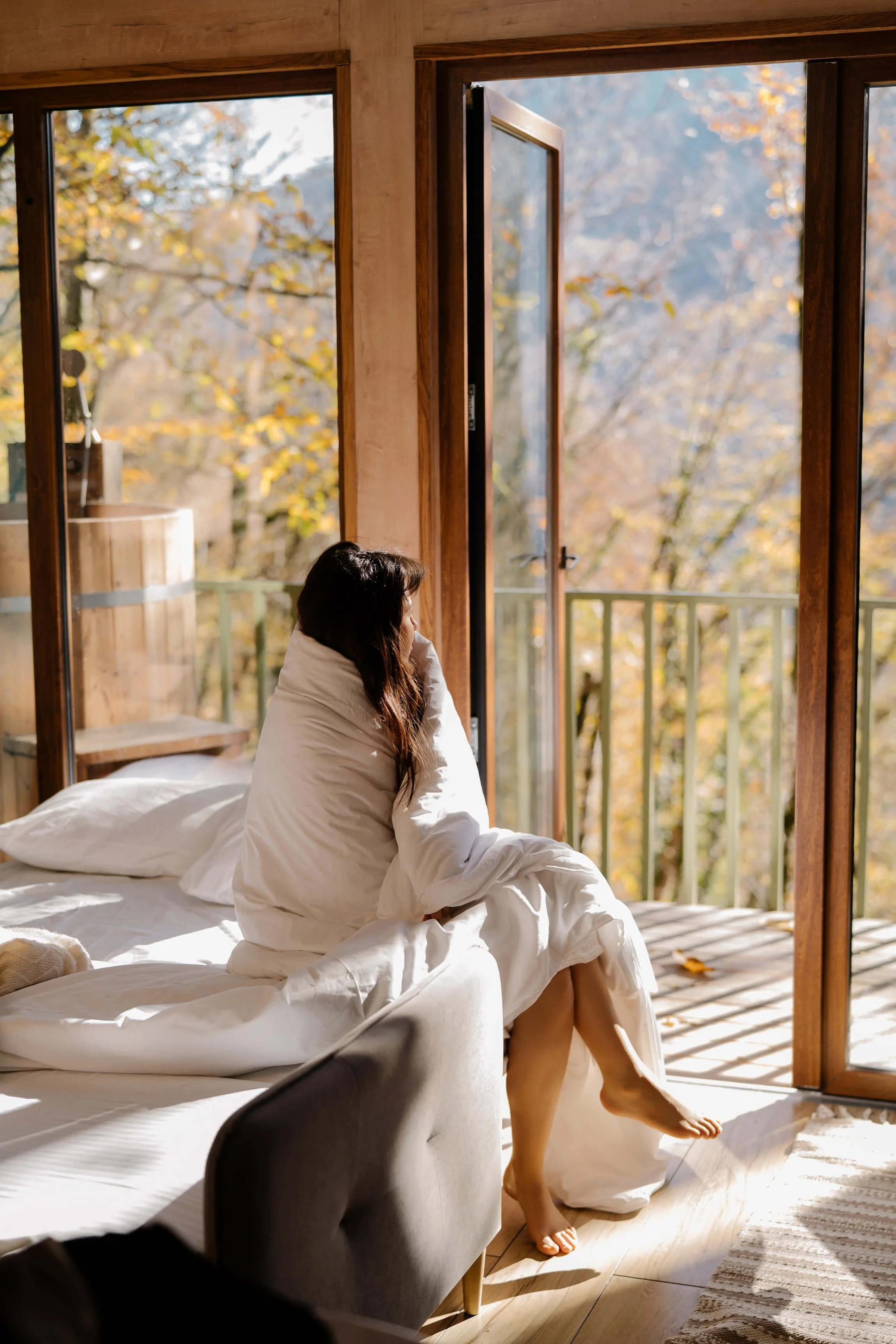 Brown haired woman sitting on her bed wrapped in a white blanket looking out her window during a fall day.