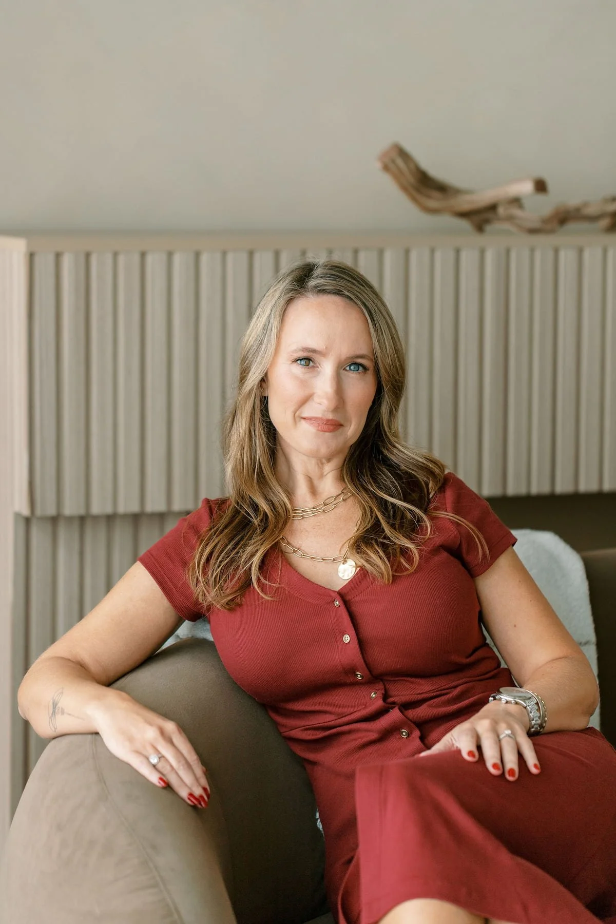 A woman with long wavy hair wearing a red dress and jewelry, sitting on a beige sofa.