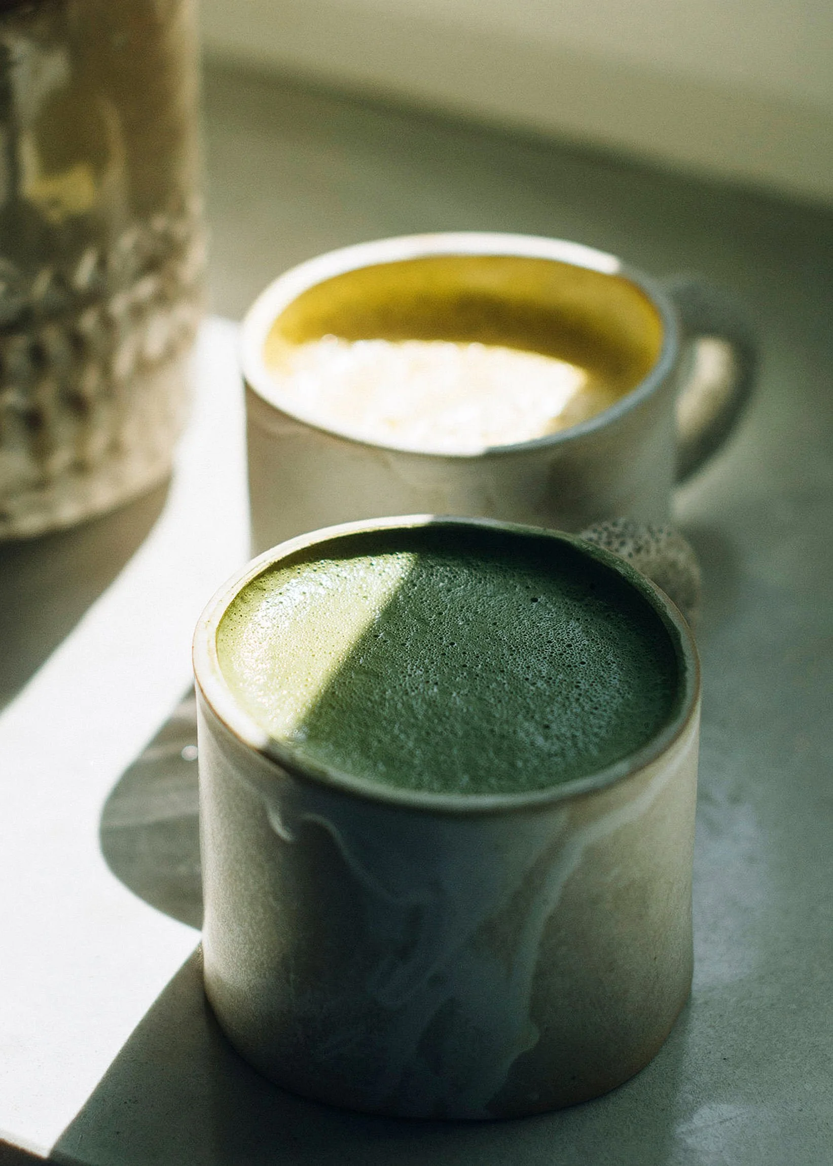 Light filtering through the window illuminating a warm cup of coffee next to a cup of matcha in ceramic mug.