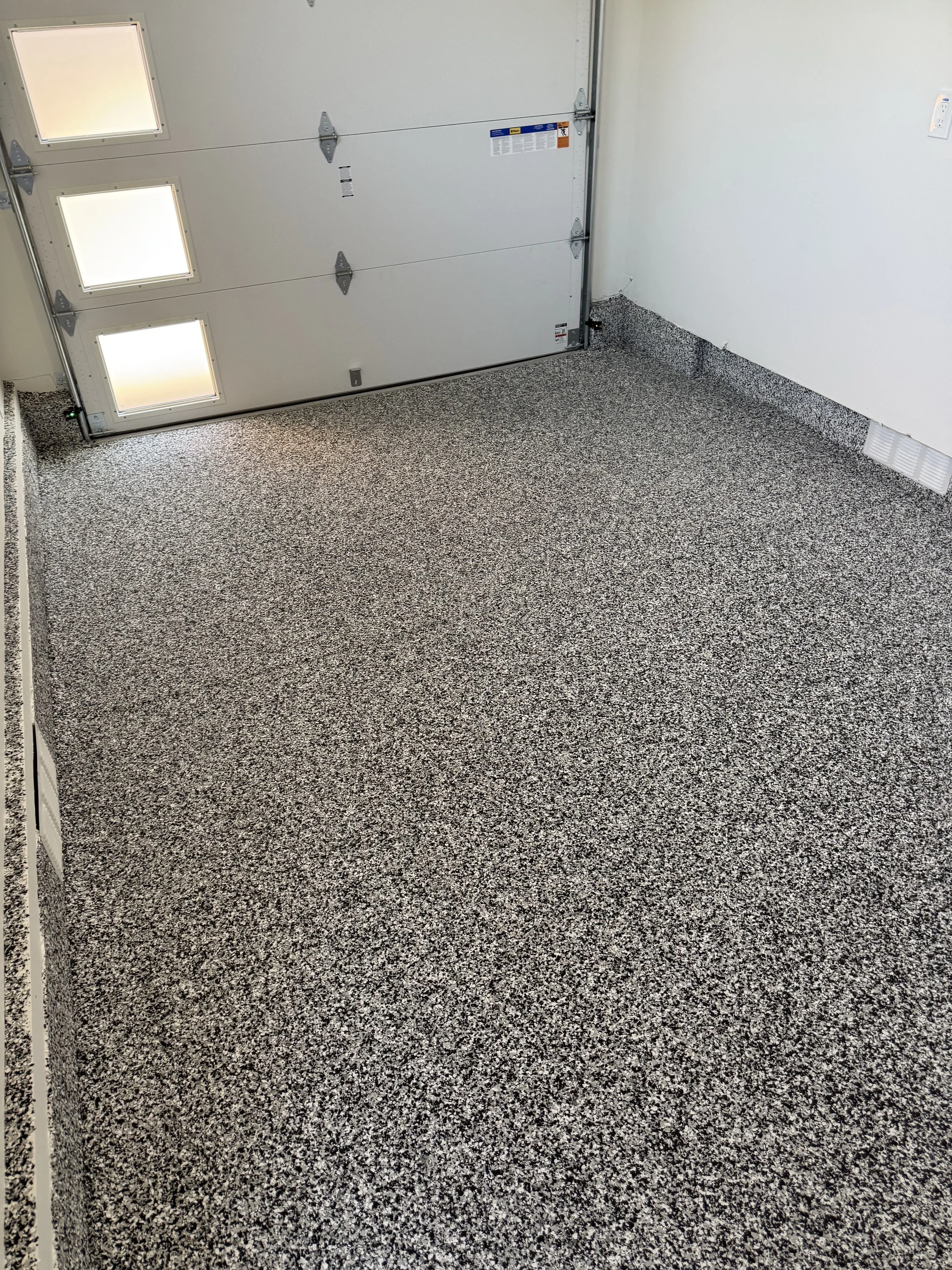 Empty garage with speckled gray and black carpet flooring, white walls, a closed garage door with windows, and a white wall vent.
