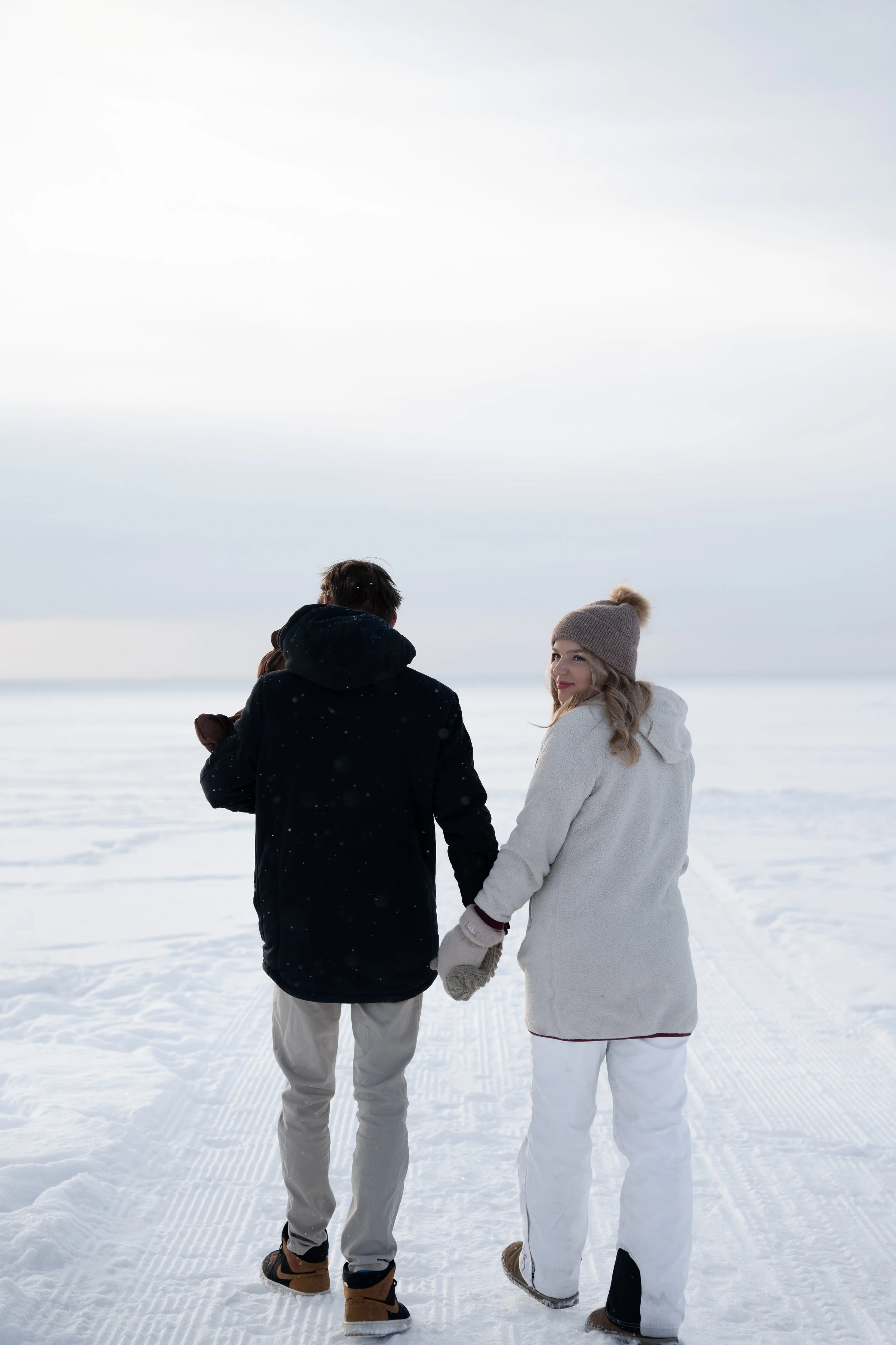 Kaksi ihmistä kävelyllä lumisella rannalla talvivaatteissa, naisen katse taaksepäin parikuvauksessa Oulussa Nallikarilla