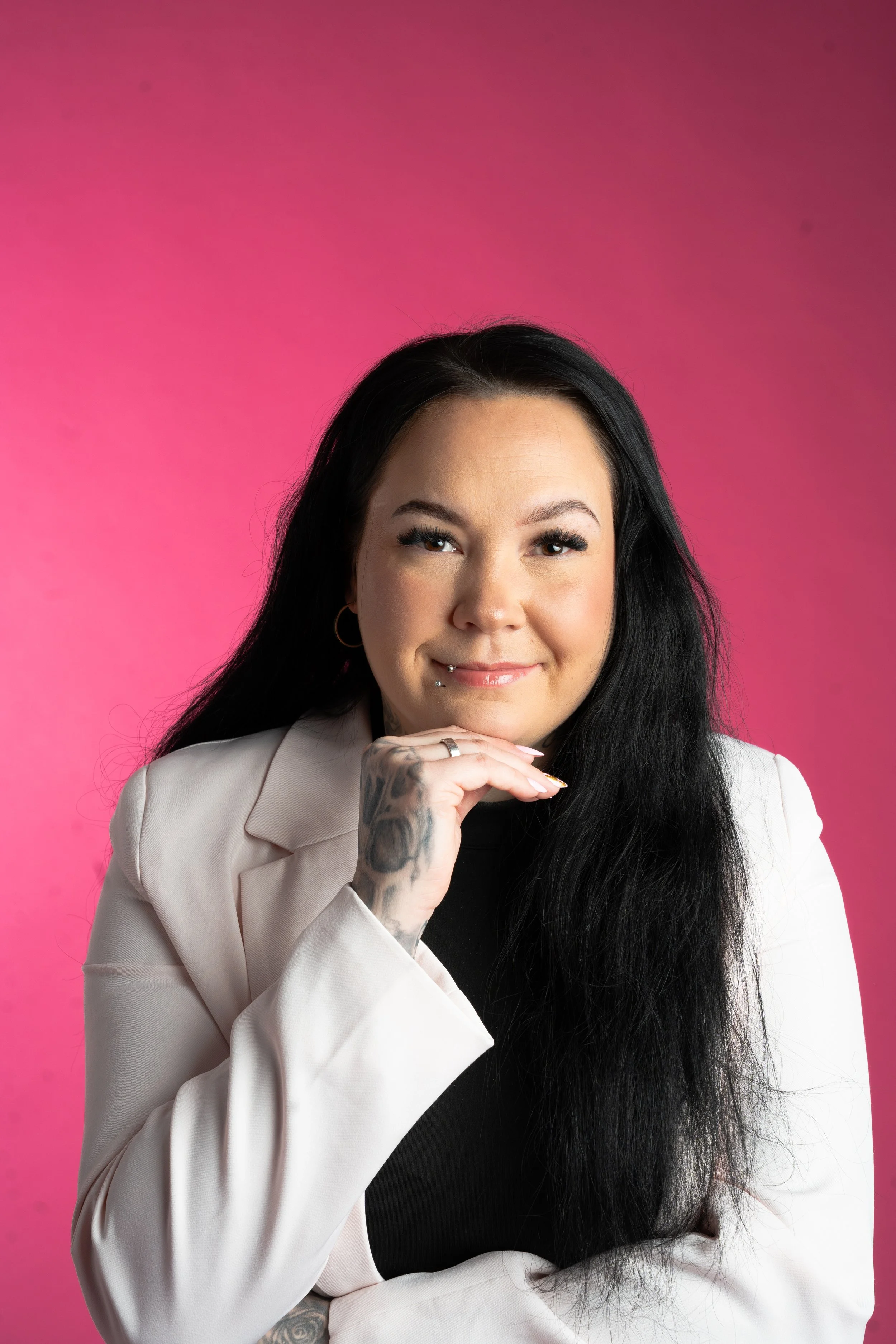 Nainen, jolla pitkä musta hiukset, hymyilee ja lepää leukansa kädellään, pinkillä taustalla.