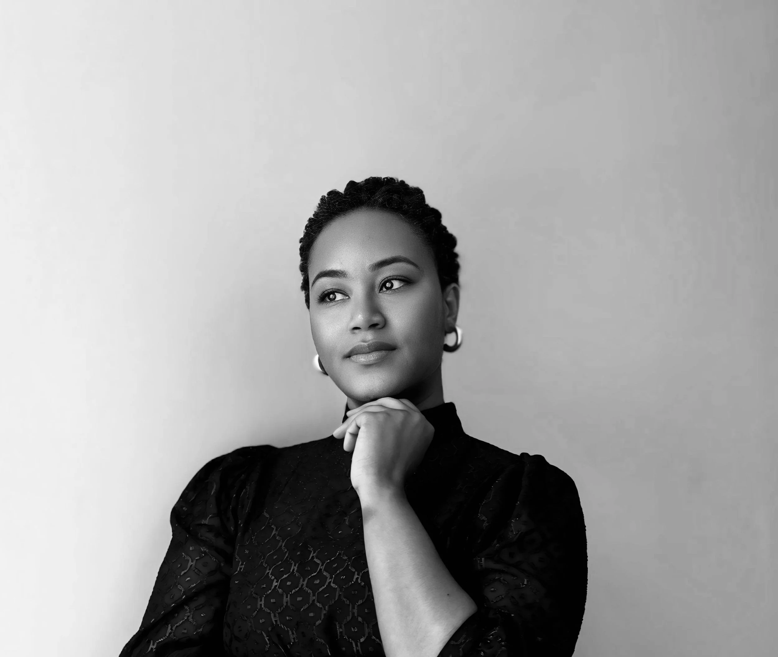 A portrait of a woman with short braided hair, wearing earrings and a dark patterned top, posed with her hand under her chin against a plain background.