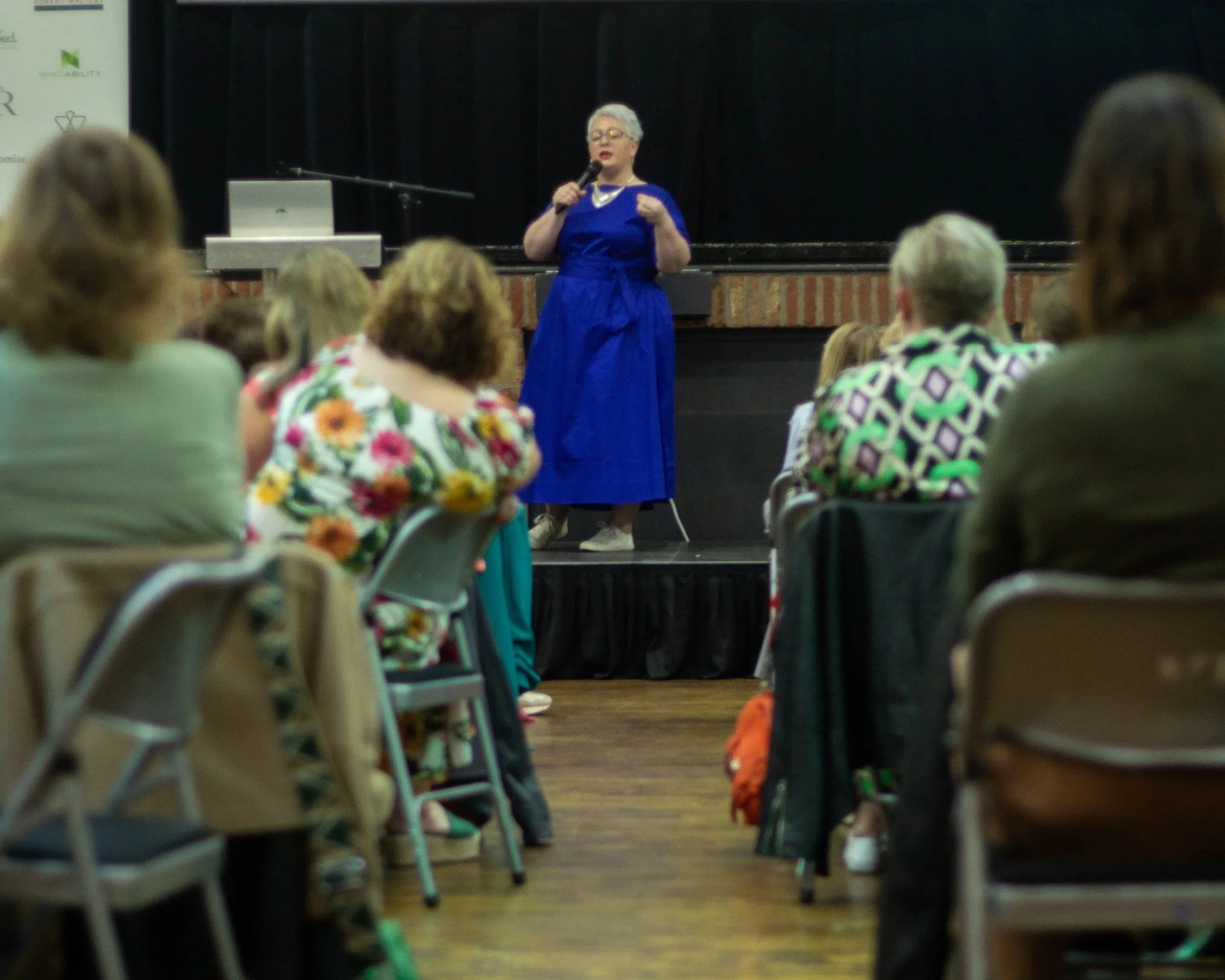 A woman with short gray hair and glasses, wearing a blue dress, is speaking into a microphone on stage at a conference or seminar. The audience members are seated and attentively listening.