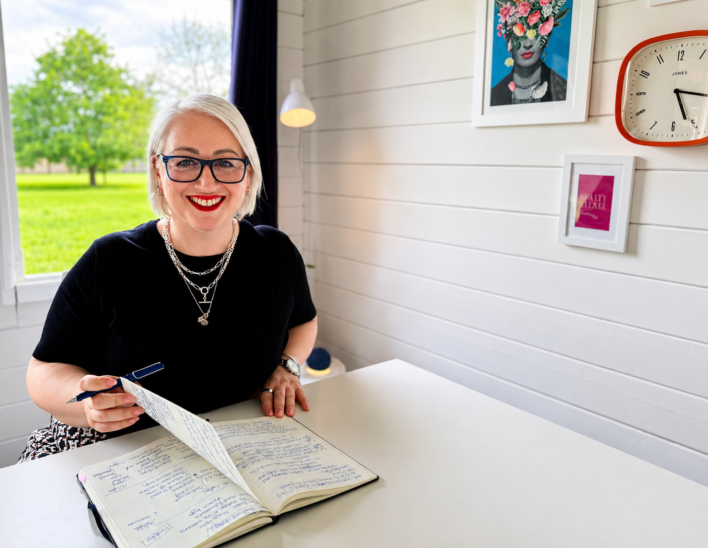 A woman with short blonde hair, glasses, and a red lipstick smile sitting at a white table with a notebook and pen, in a bright room with white wall paneling, framed wall art, and a clock, with a window showing a green outdoor scene.