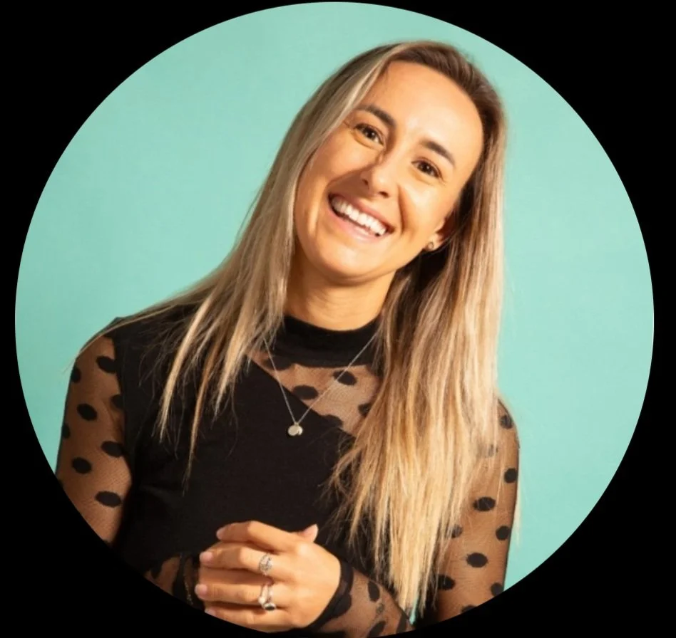 A woman with long, blonde hair smiling at the camera, wearing a black top with sheer, polka-dotted sleeves and a necklace, standing against a teal background.