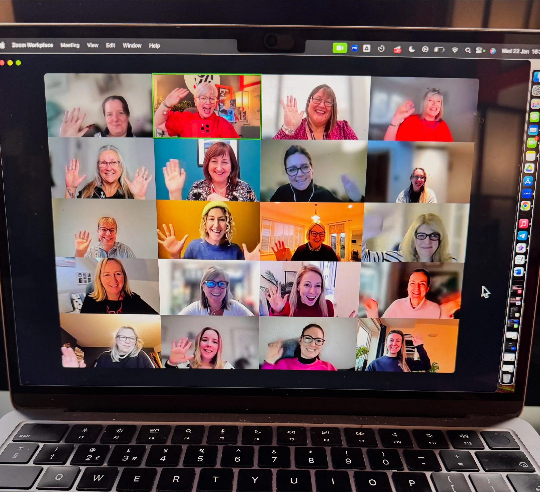 Laptop screen showing a virtual meeting with 20 smiling women waving and celebrating, in a grid of video call windows.