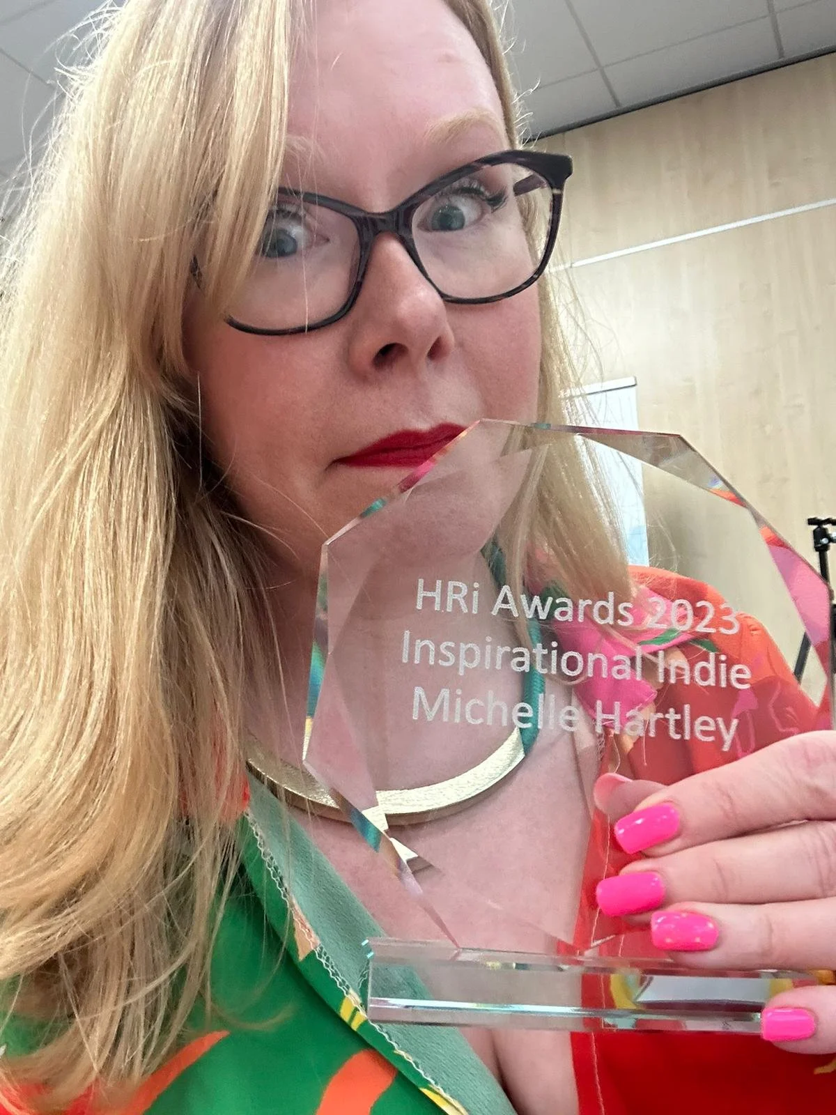 A woman with glasses and red lipstick holding a transparent award with the inscription 'HRi Awards 2023 Inspirational Indie Michelle Hartley'