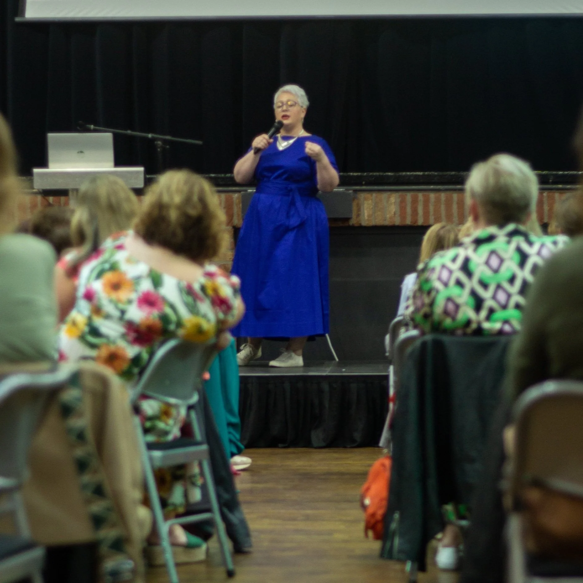 A woman with short gray hair and glasses is speaking into a microphone on a stage with a brick wall backdrop during an event or presentation. She is wearing a long blue dress.
