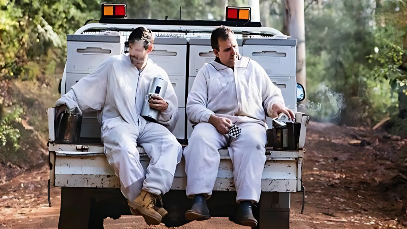 Two men in white beekeeping suits sitting on the back of a pickup truck in a wooded area, smoking and drinking coffee.