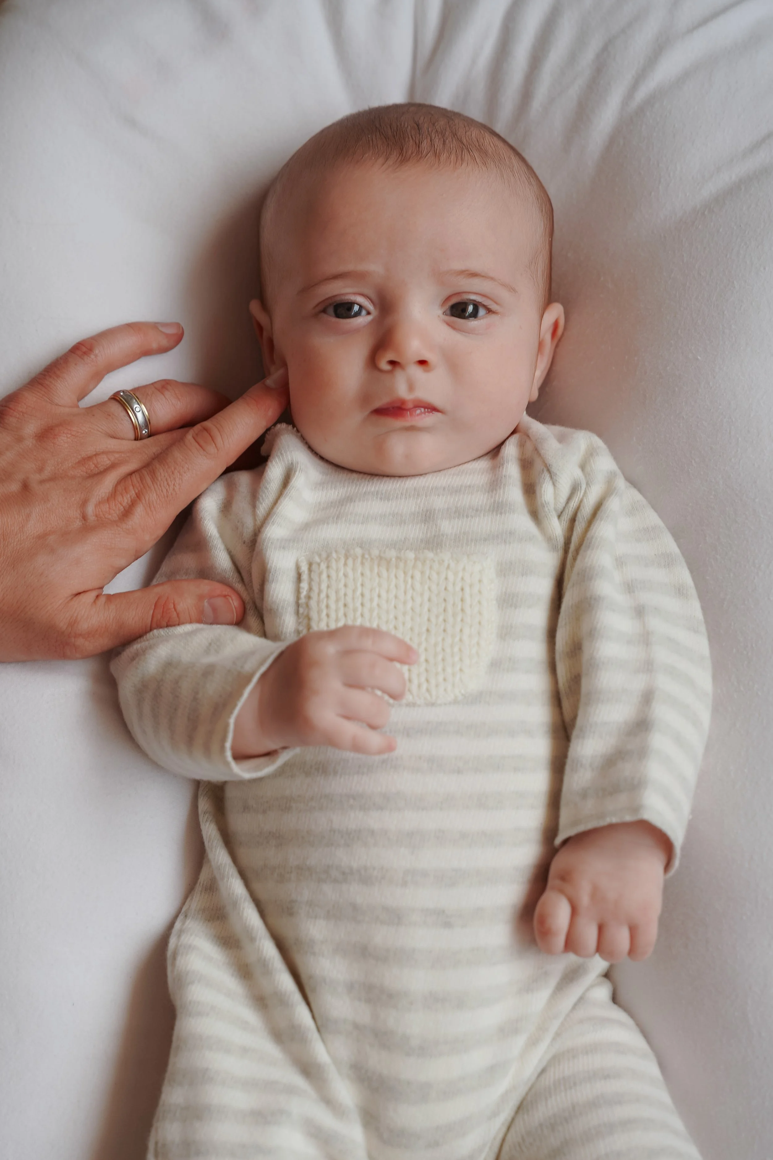 Close-up of a baby with light brown hair lying on a white surface, wearing a cream and beige striped outfit, with a hand touching the baby's cheek.