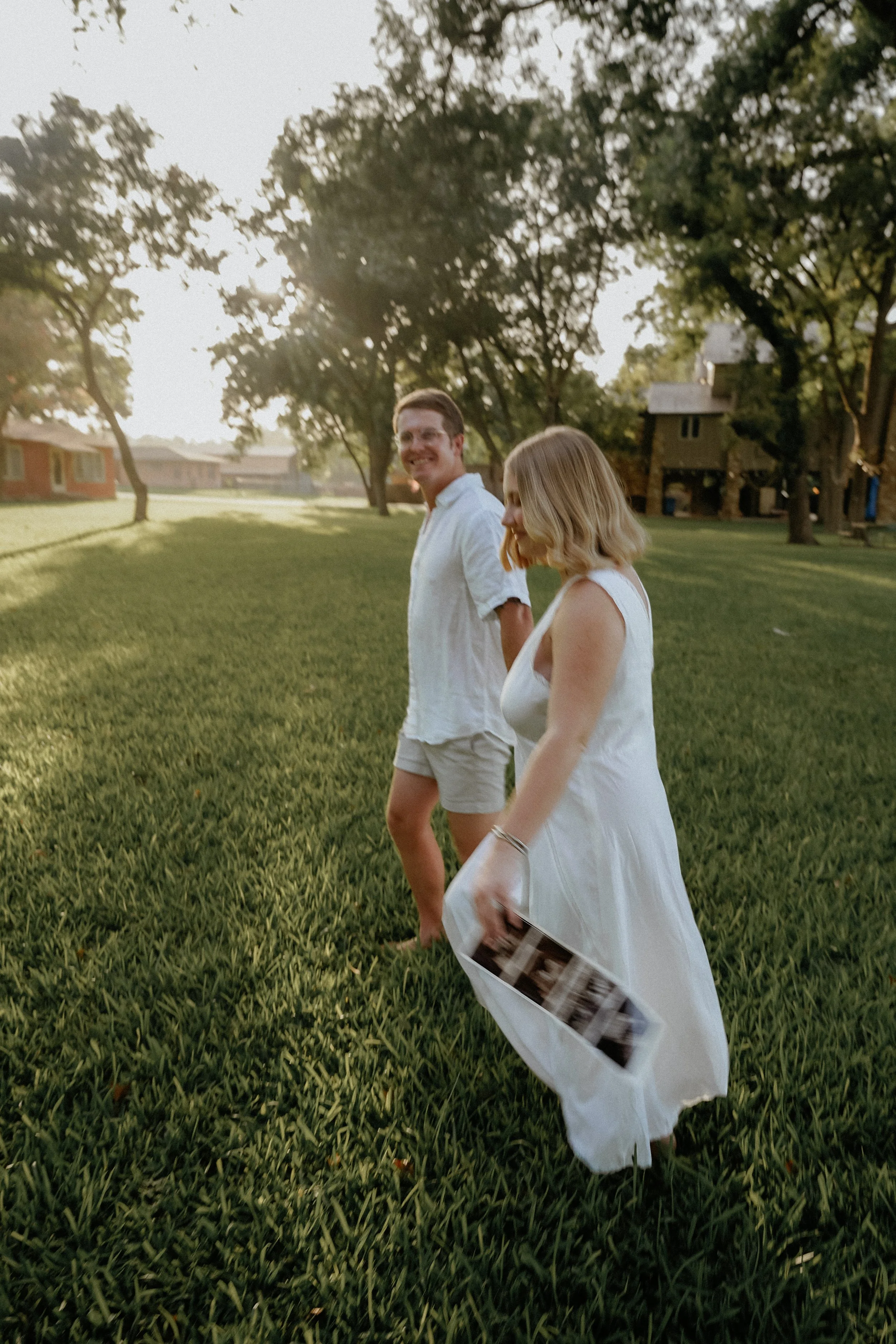A man and a woman walking on a grassy field with trees and houses in the background during sunset, both smiling and dressed in white clothing. The woman is holding photographs. Lake Dunlap maternity session at golden hour with ultrasound photo
