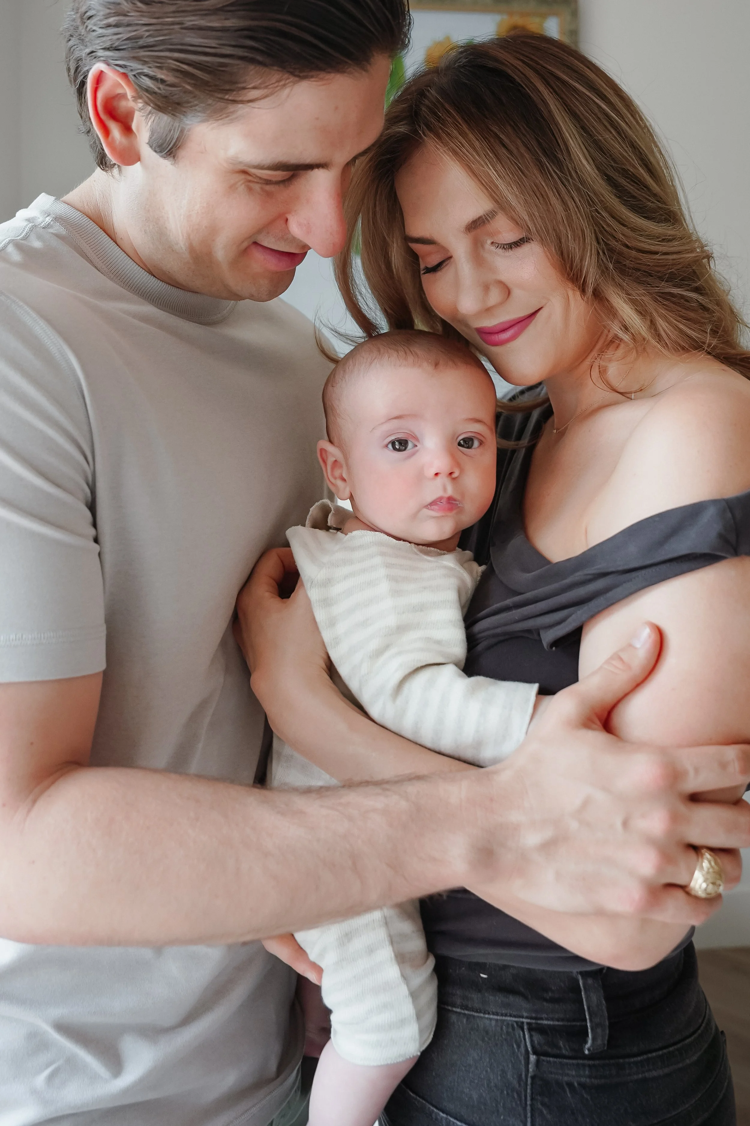 A family of three, including a man, a woman, and a baby, embracing each other close while smiling and looking at the camera. Lifestyle mother and baby photographer session. In home family photography in Austin and New Braunfels Texas.