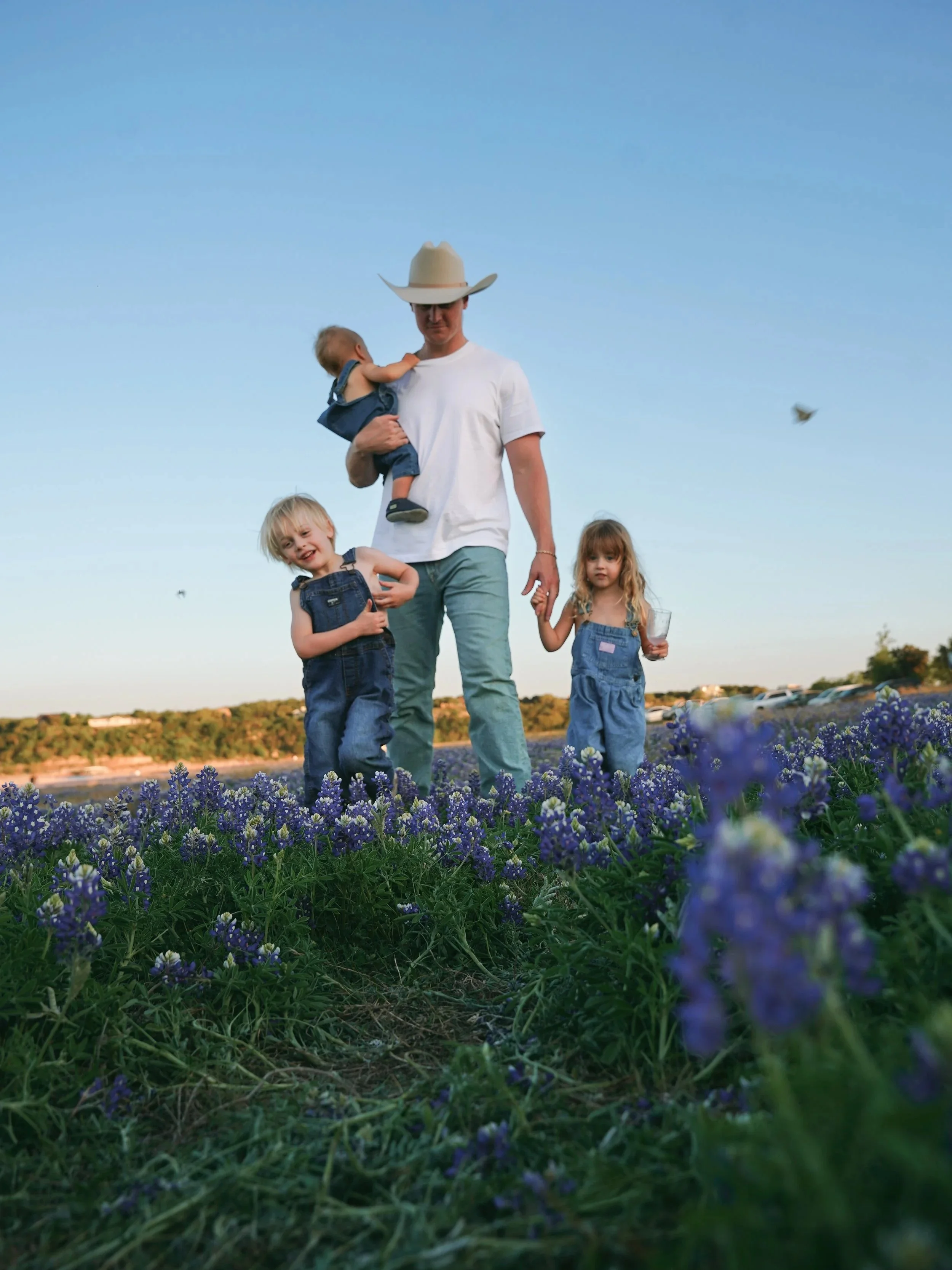 A man wearing a cowboy hat and white T-shirt holding a small child in blue overalls, walking through a field of purple flowers with two young children, one smiling and one holding a glass, against a clear blue sky. Bluebonnet family session in Texas.