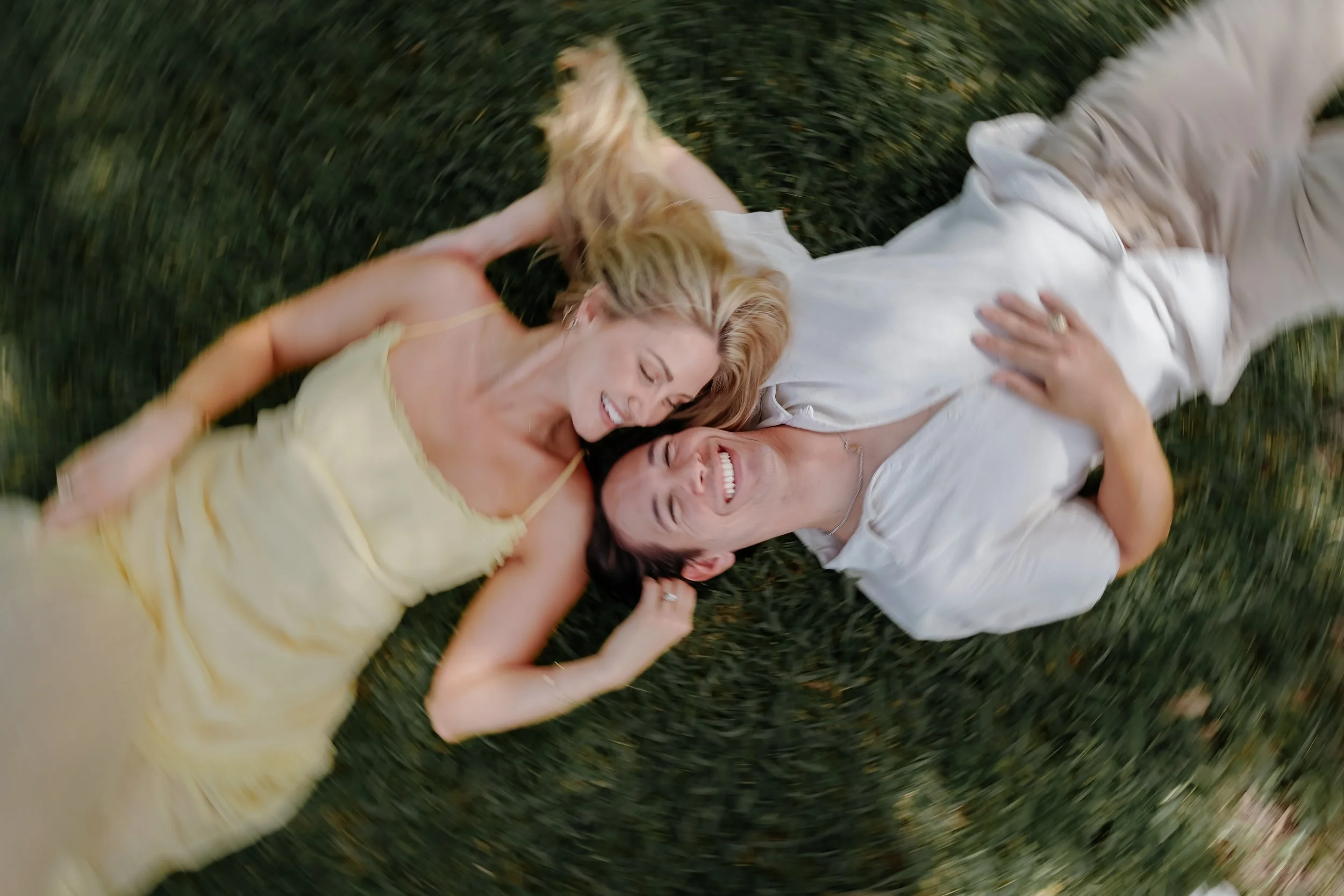 Two people lying on grass, smiling and hugging each other, enjoying outdoors. Engagement session where the camera is swirling and spinning. Couples photoshoot at the Contemporary Austin Laguna Gloria.