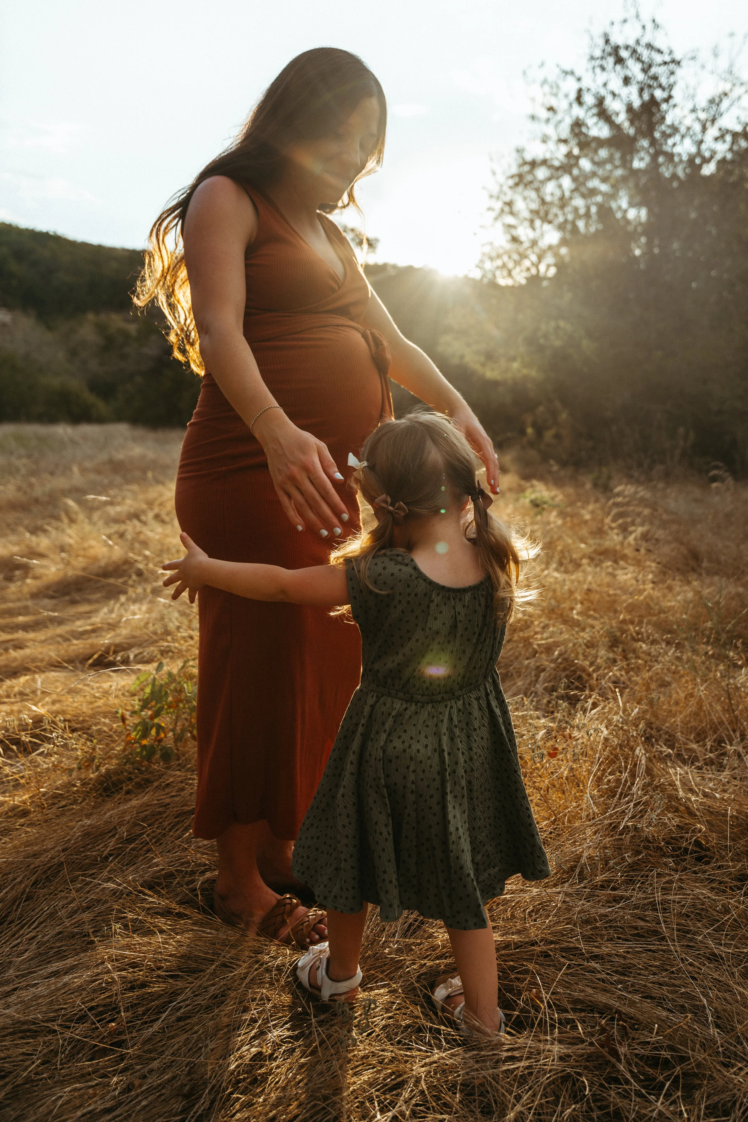 Austin Portrait Photographer. A pregnant woman in a brown dress standing in a field of dry grass with a young girl, back to the camera, with pigtails, both facing the sunset.