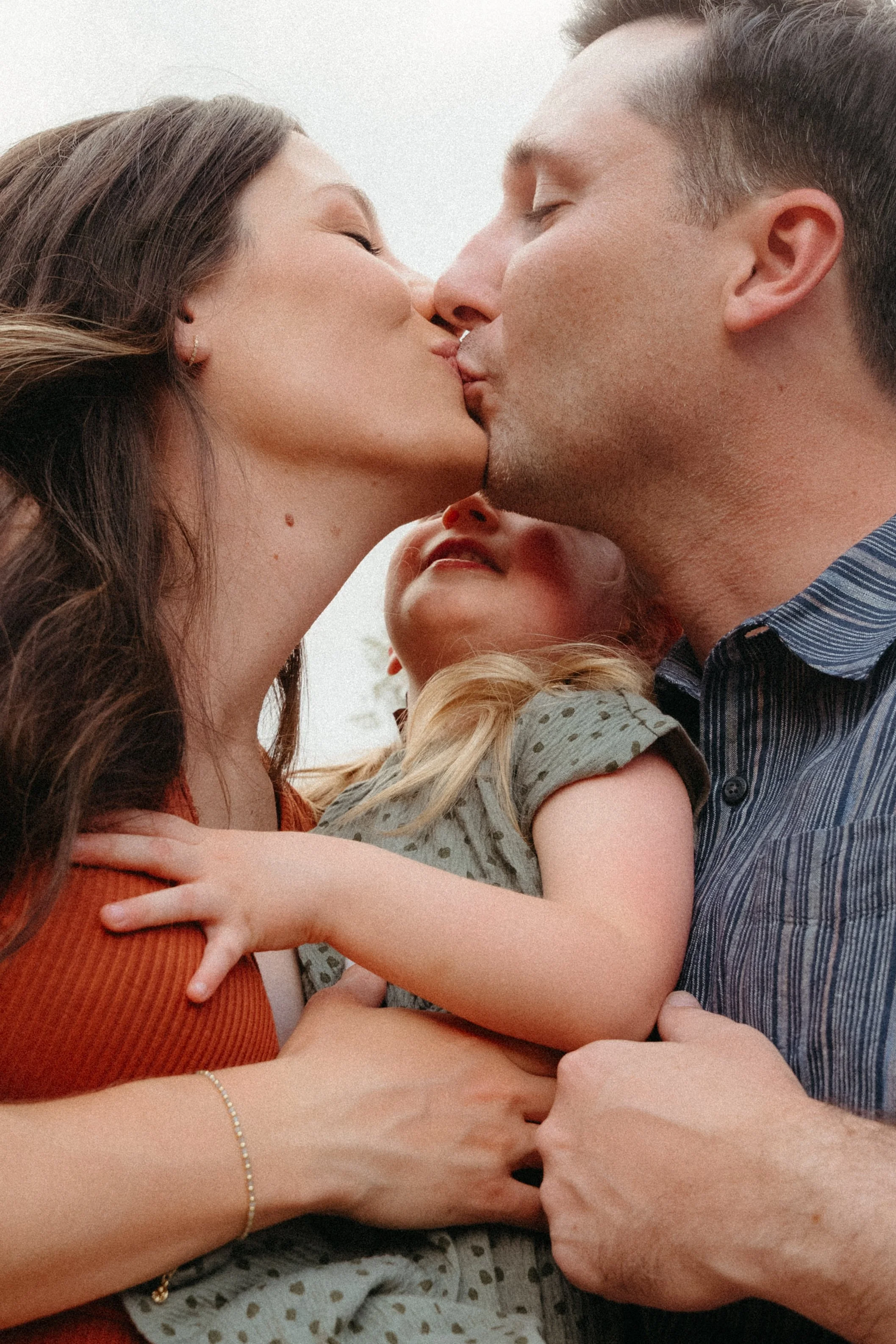 Austin Portrait Photographer. A couple kisses while holding a young girl between them. The woman has long brown hair and the man has short brown hair. The girl has blonde hair and is wearing a polka-dotted shirt.