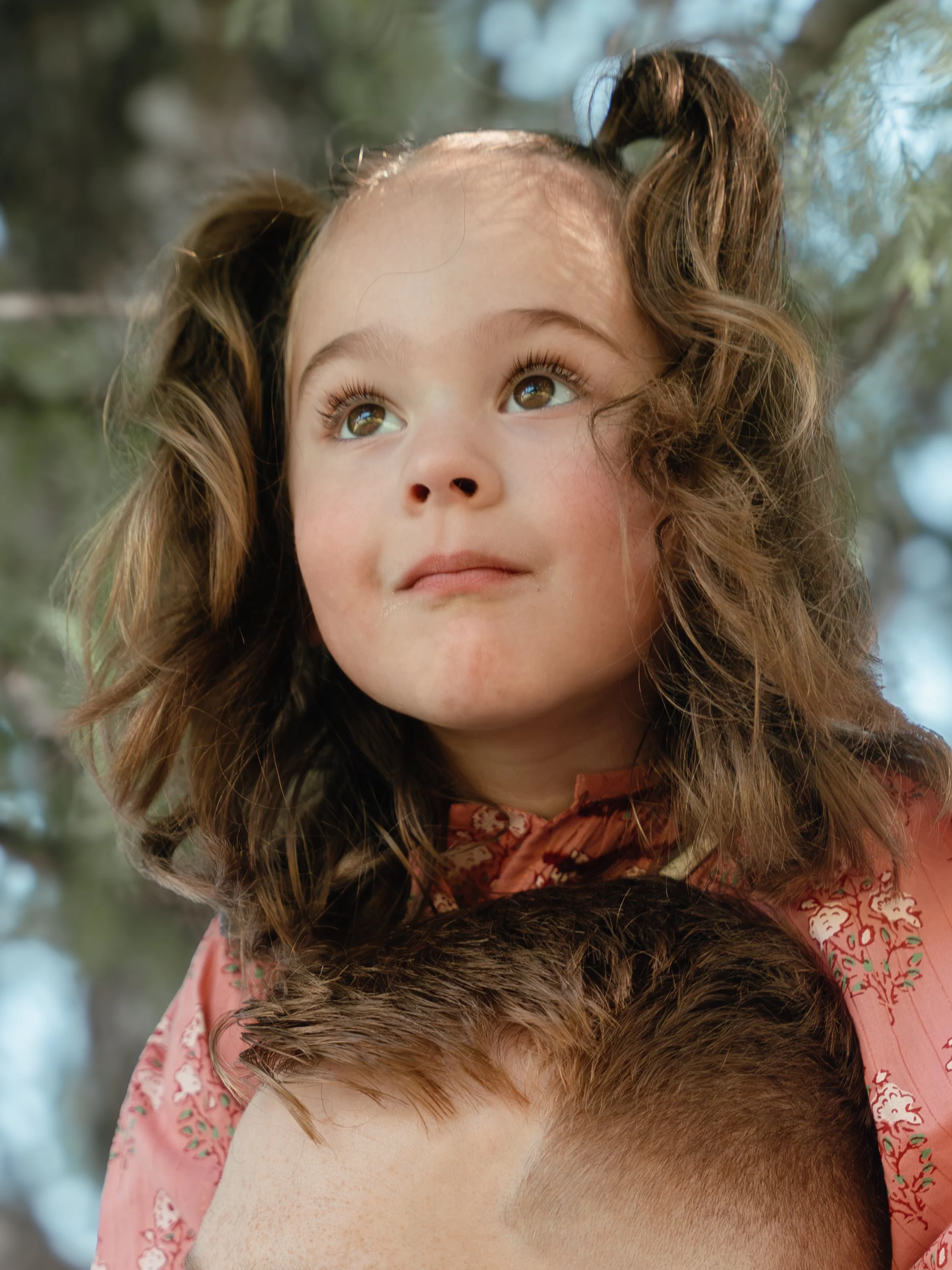 Austin Family Photographer/ A young girl with curly brown hair, wearing a pink patterned shirt, looking upward outdoors with a background of blurred trees.