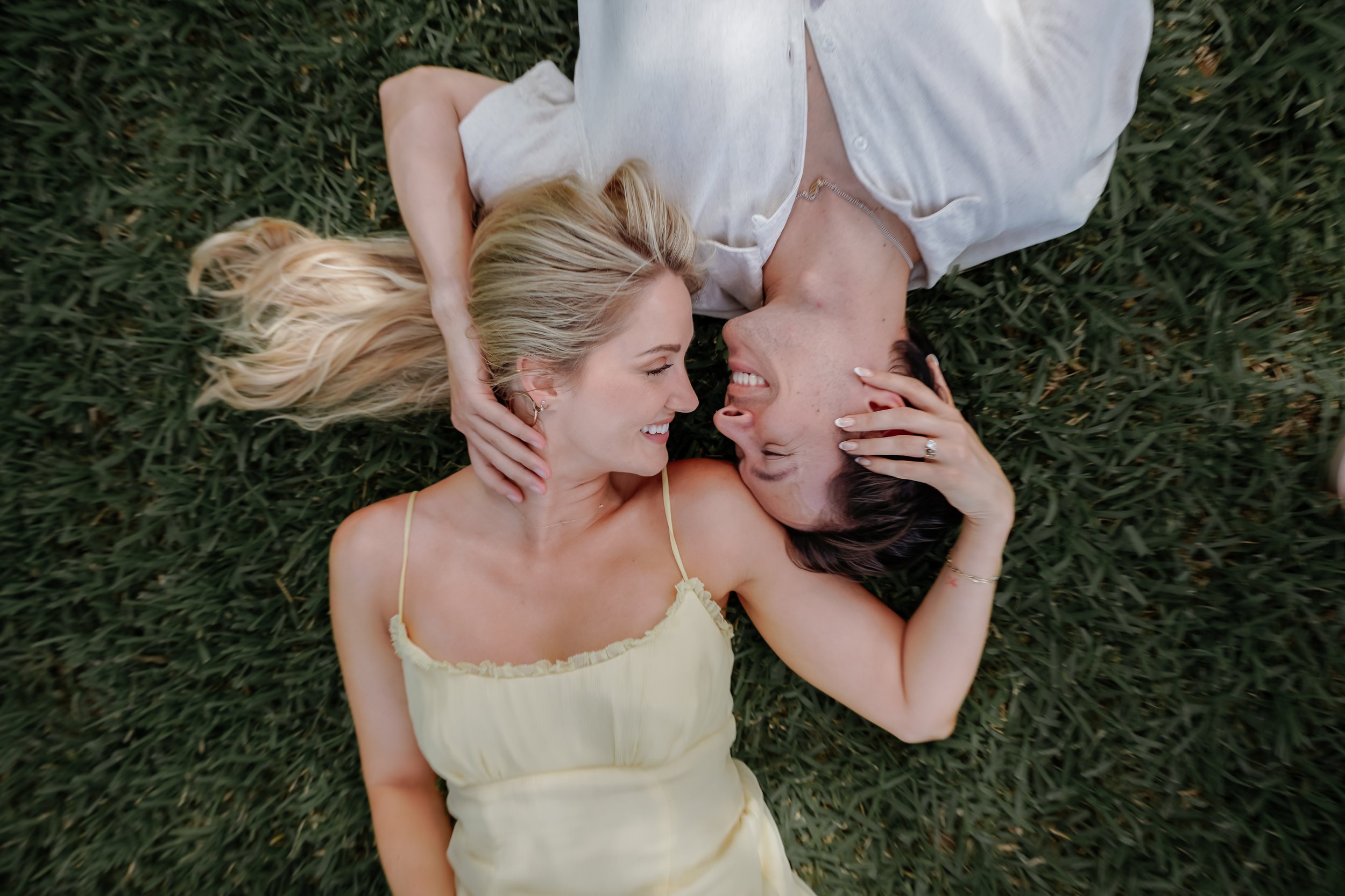 Two people lying on the grass, facing each other and smiling, with one woman wearing a light yellow dress and the other in a white top, engaging in a close, happy moment outdoors. Engagement photograph session at Laguna Gloria at the Contemporary
