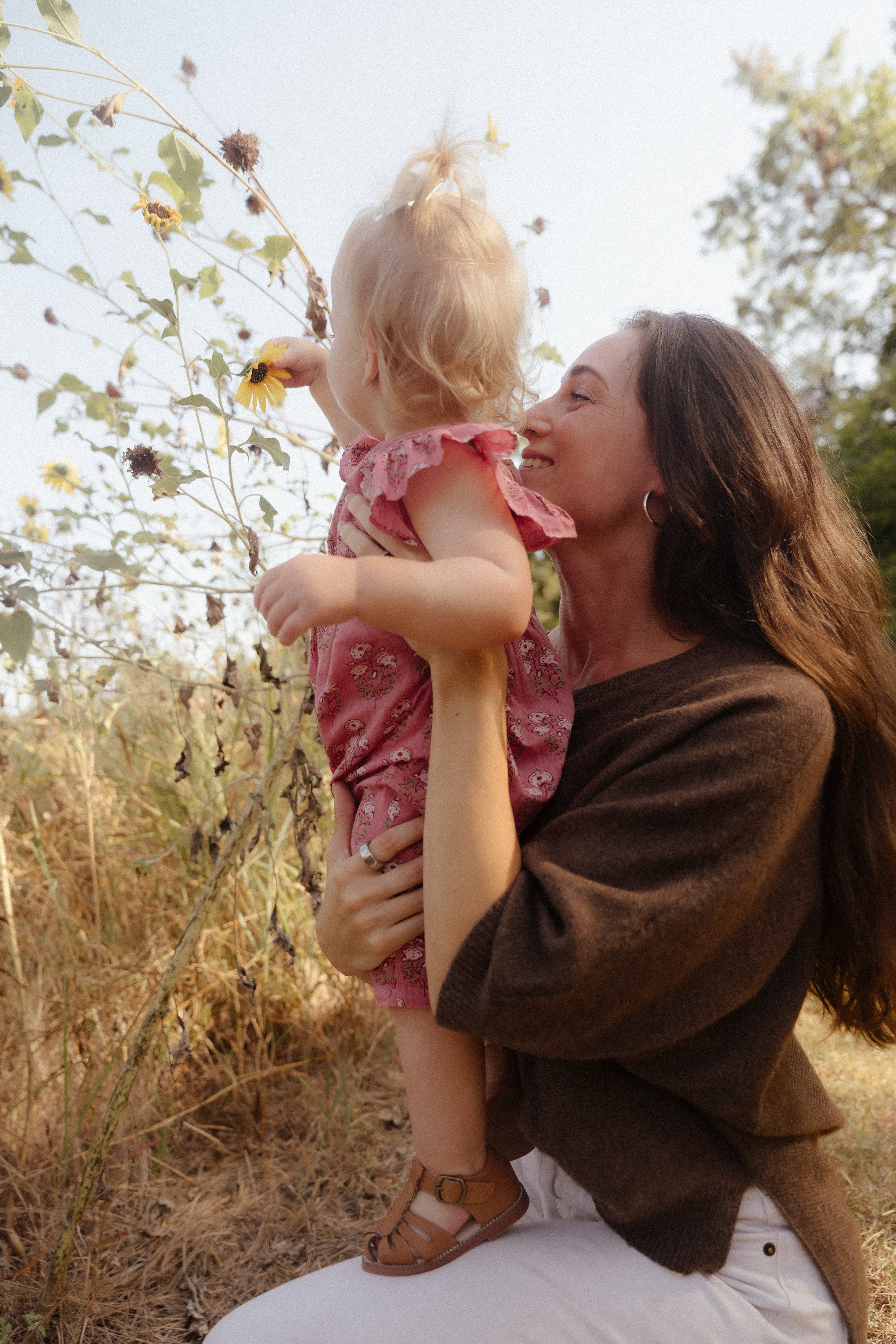 A woman holding a young girl outdoors, both smiling, with the girl touching a sunflower on a nearby plant.