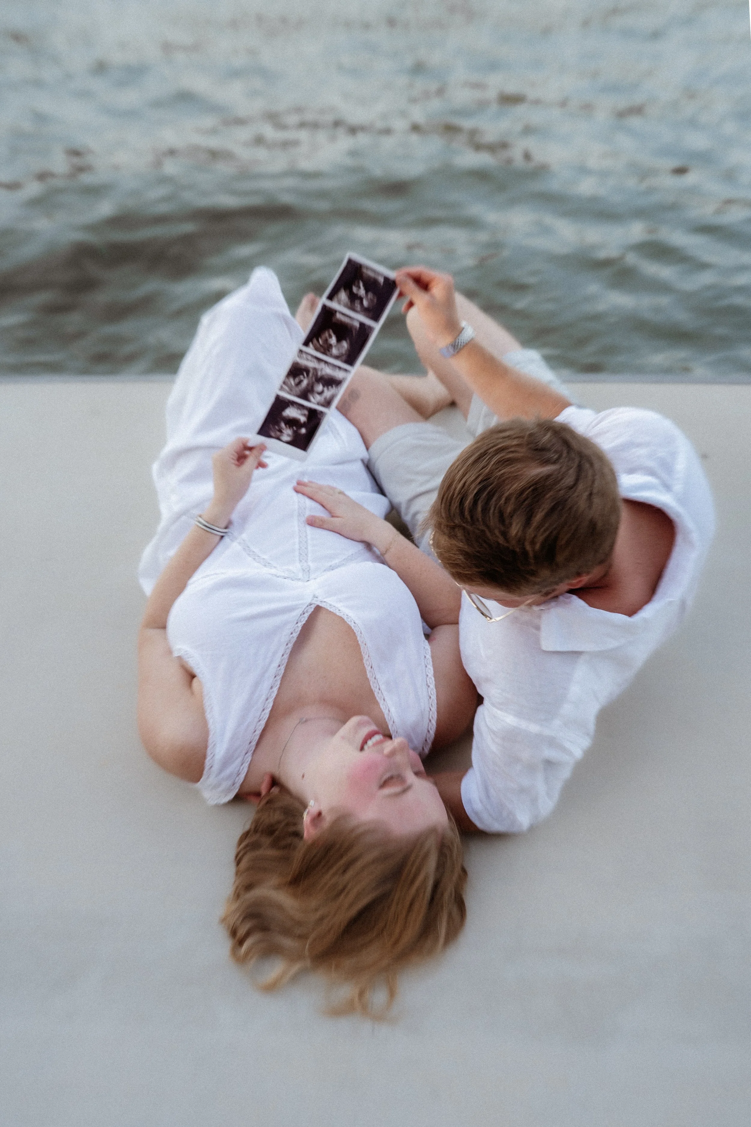 A woman and a man lie on the ground by water, holding ultrasound images, with the woman resting her head on the man's shoulder. Maternity session where the couple is on a dock by the lake in New Braunfels on Lake Dunlap holding a sonogram ultrasound.