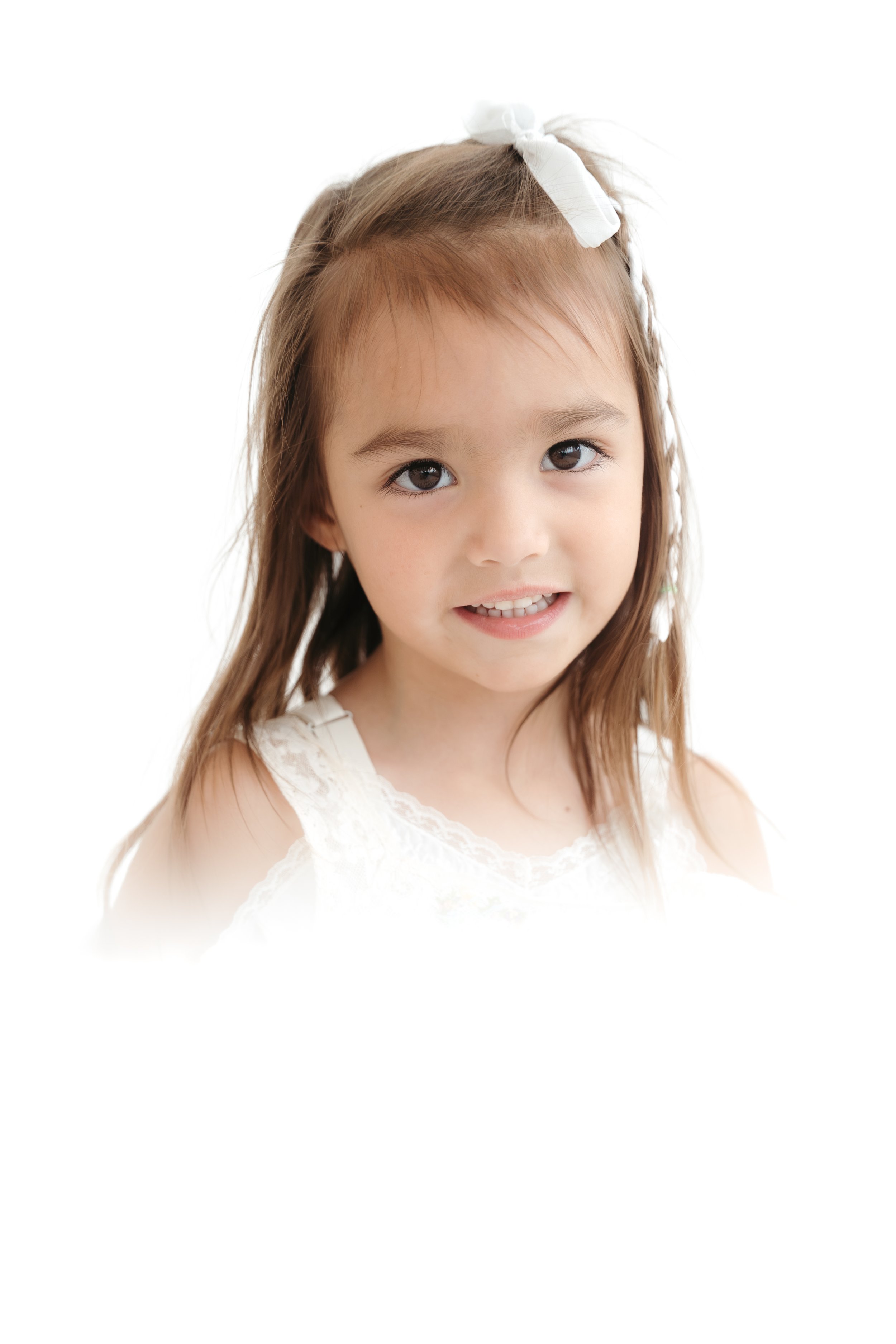 Close-up of a young girl with brown hair, wearing a white dress and a white hair bow, smiling softly against a plain white background.