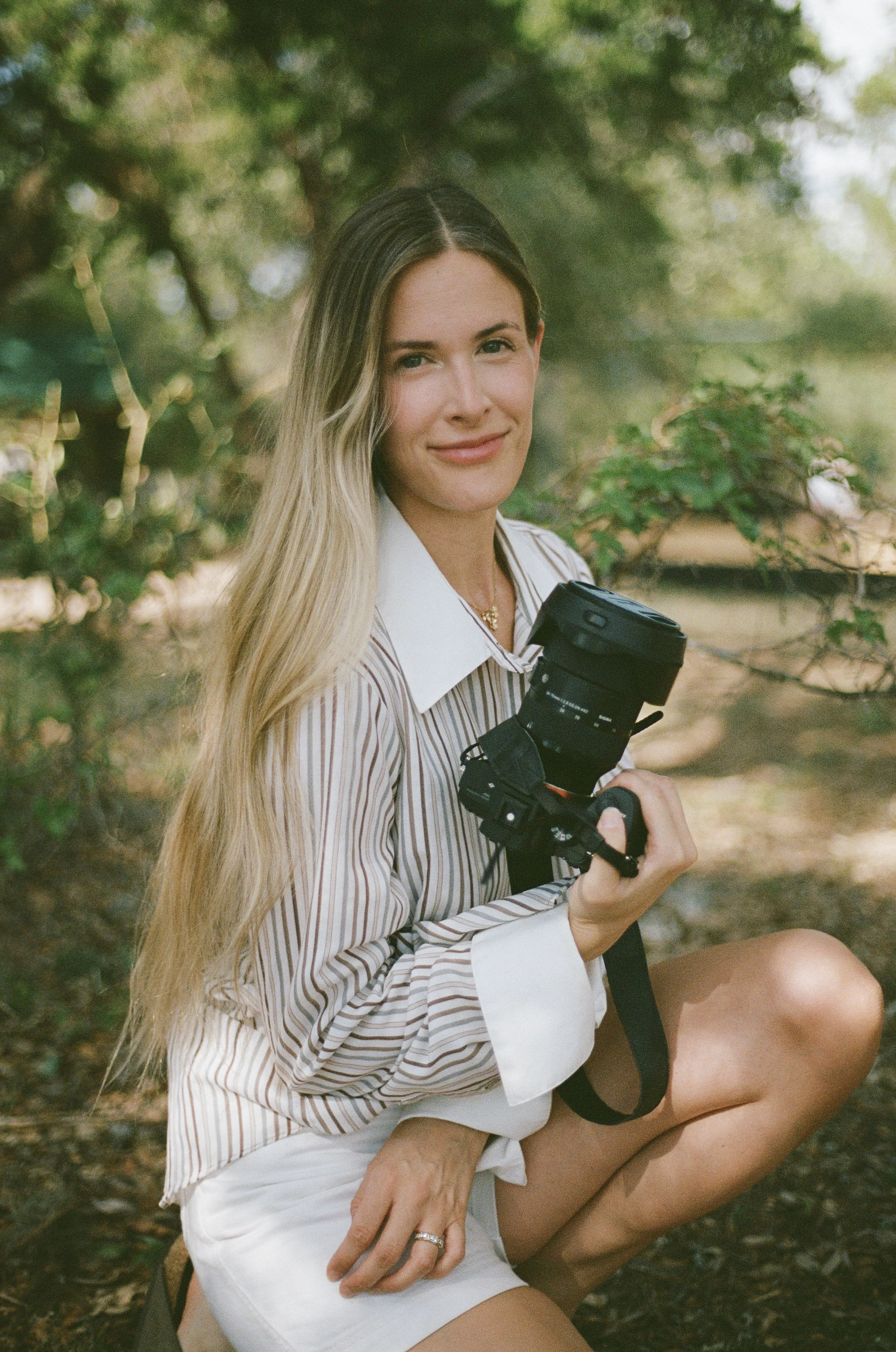 Austin Portrait Photographer. A woman sitting outdoors on grass, holding a camera, with long blonde hair, wearing a striped shirt and white shorts, smiling at the camera, surrounded by green trees.