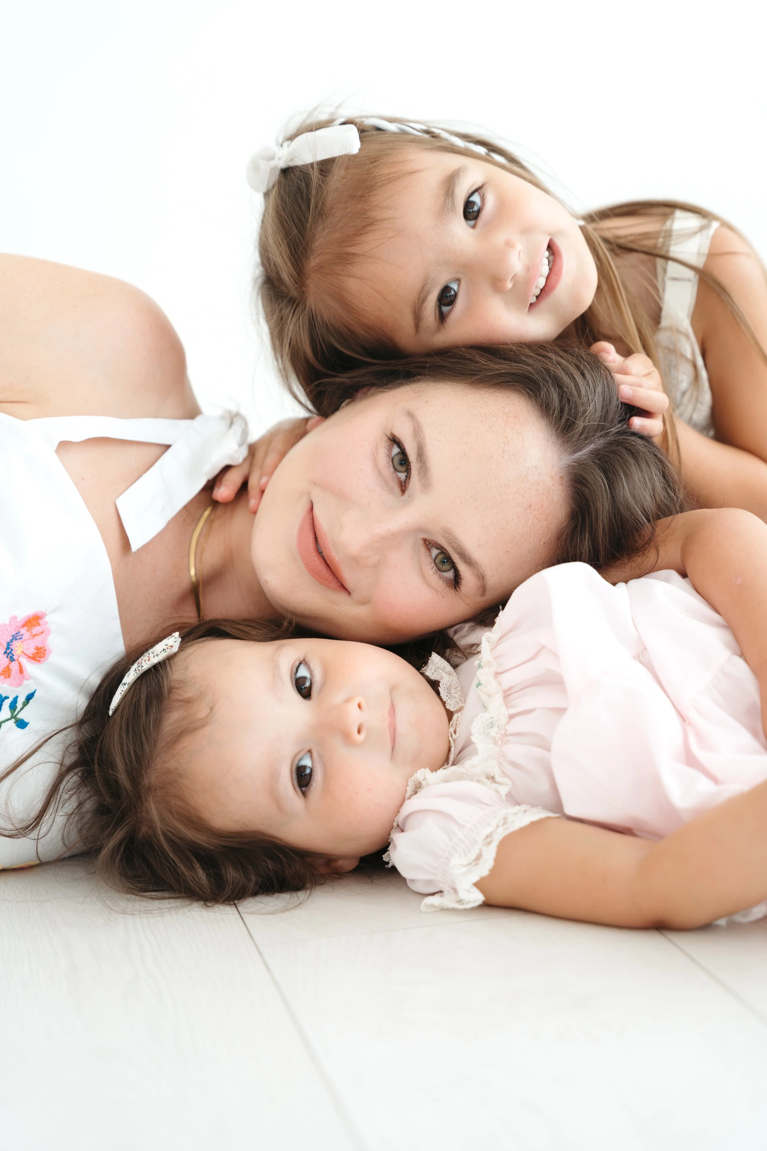 A woman lying on the floor with four young girls cuddling and resting their heads on her. All appear relaxed and happy, with the girls smiling softly. The background is white. Heads stacked portrait photography in Photography Studio in Austin Texas