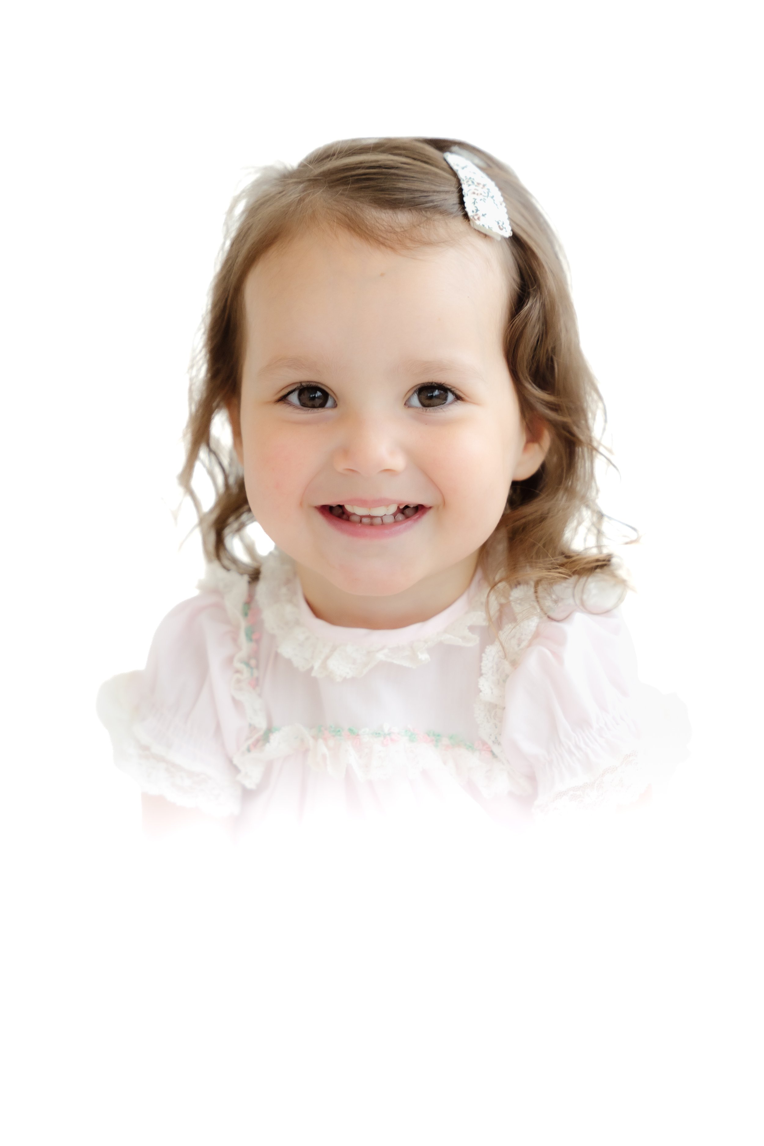 A smiling young girl with light brown hair, wearing a light pink dress with lace details and a white bow hairclip, against a white background. Heirloom white background diffuse cloud-like vintage inspired photograph.