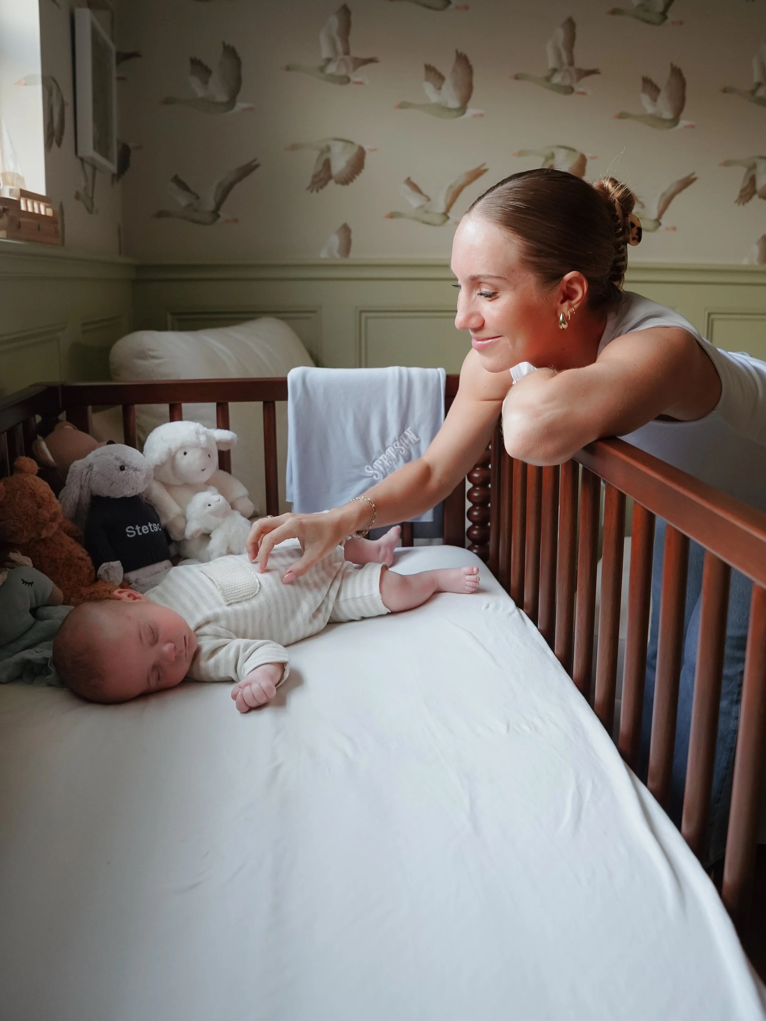 A woman leaning over a crib with stuffed animals and a baby sleeping on a bed. In home family photography lifestyle session in Texas.